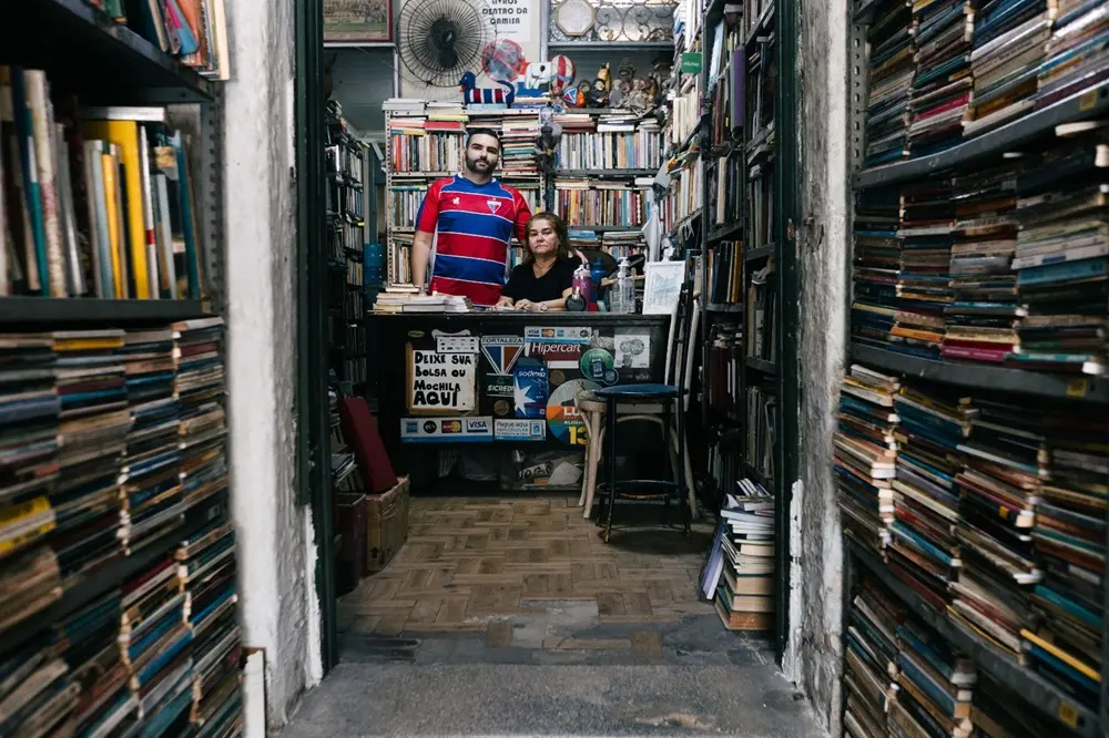 Na imagem, uma tomada em plano médio, através de uma porta ou corredor estreito, que mostra o interior de um sebo (livraria de livros usados). As laterais da imagem são dominadas por estantes altas e abarrotadas de livros, criando um túnel visual. No centro, ao fundo, atrás de um balcão, estão duas pessoas: um homem e uma mulher. O homem, em pé e ligeiramente atrás, usa uma camisa polo azul e vermelha. A mulher, sentada ou inclinada, usa uma blusa preta. Ambos olham diretamente para a câmera. O balcão à frente deles tem adesivos de cartões de crédito e um cartaz que diz, em parte, 