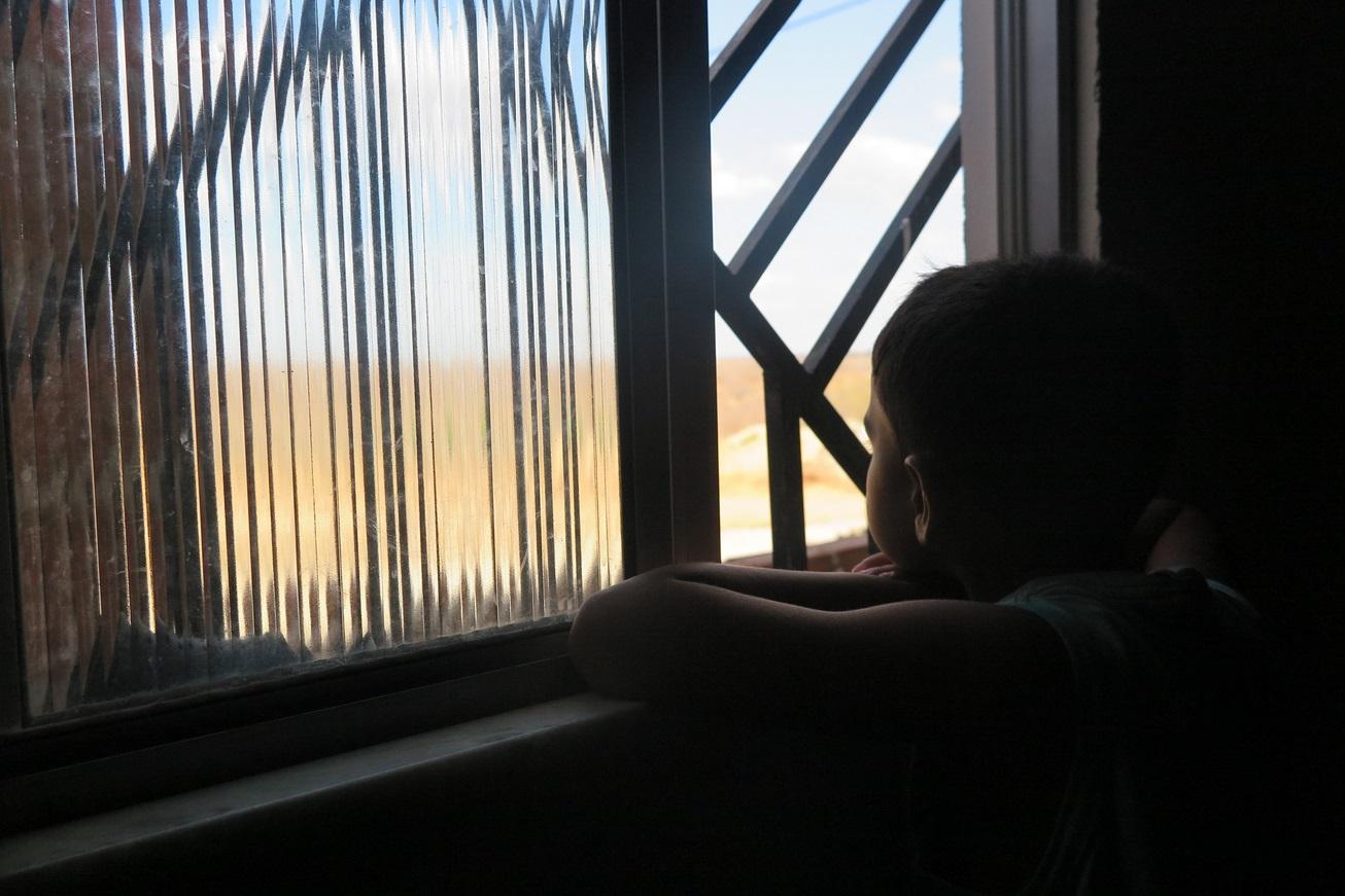 Silhueta de menino olhando por janela com grades e vidros. Ele apoia os braços no parapeito. A cena demonstra isolamento e solidão.