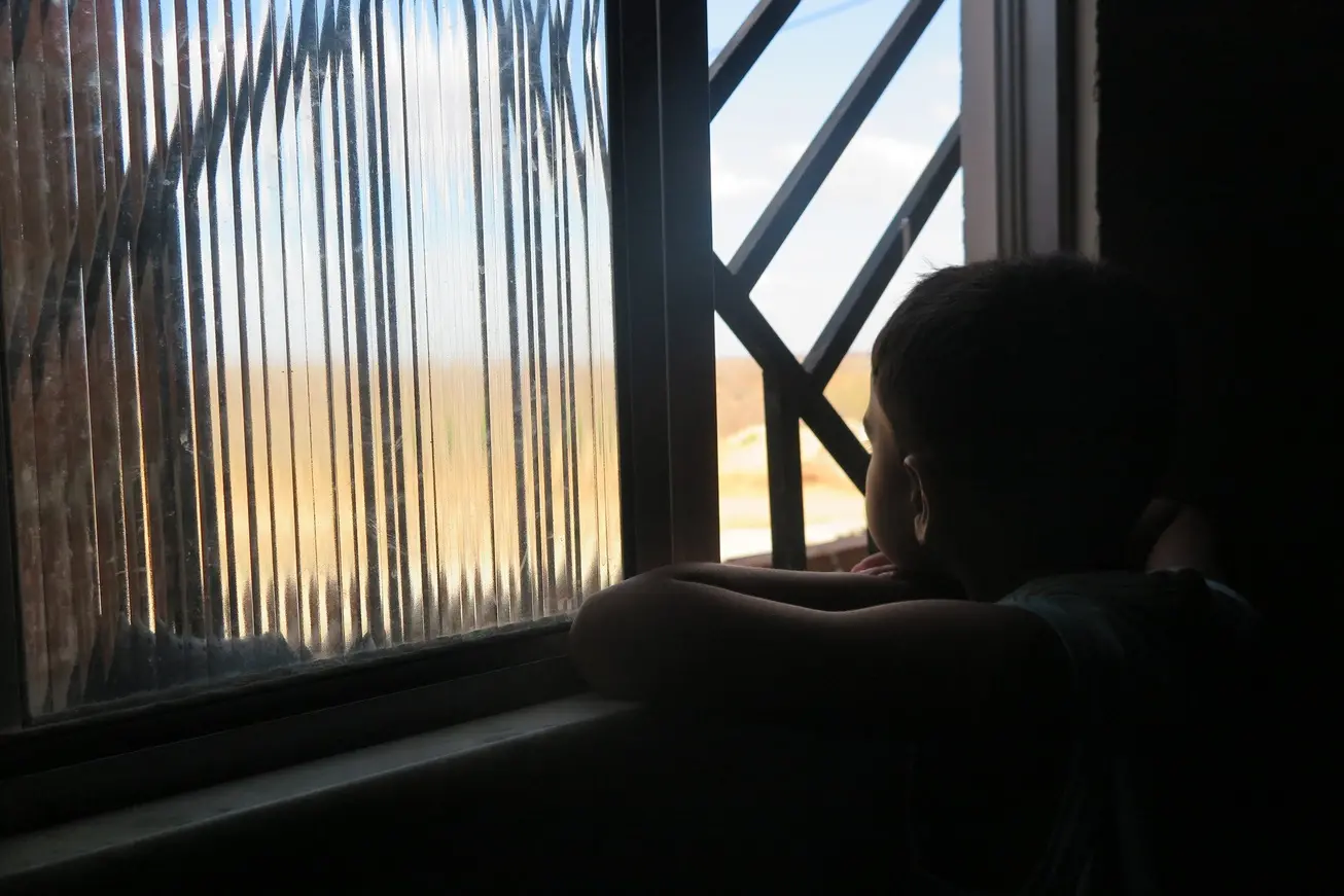 Silhueta de menino olhando por janela com grades e vidros. Ele apoia os braços no parapeito. A cena demonstra isolamento e solidão.