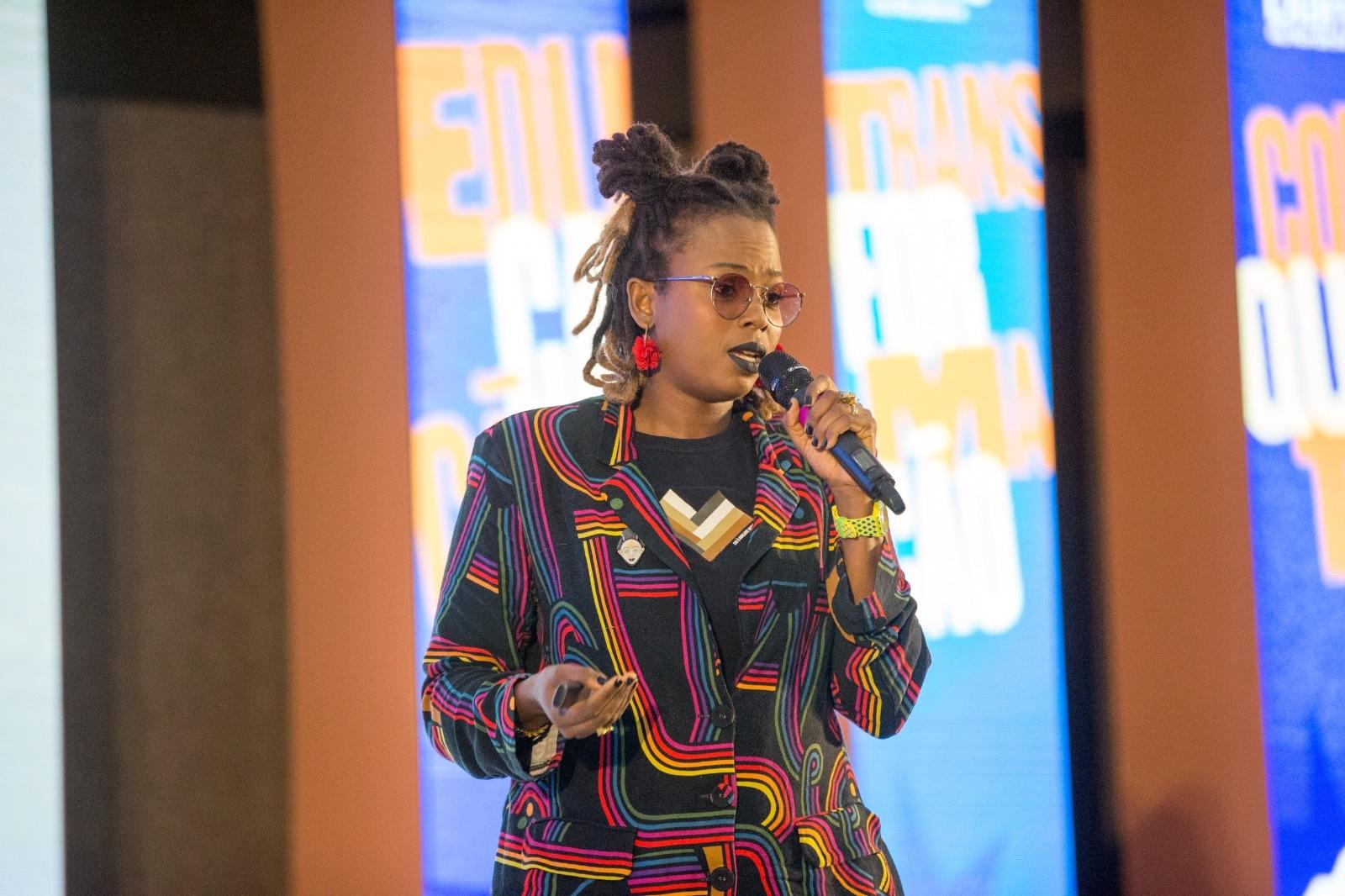 Imagem mostra Luana Genot, mulher preta de cabelo crespo, com microfone na mão, óculos de lentes redondas, roupa preta com estampas coloridas e brincos de flores vermelhas. Ela está palestrando em um palco.