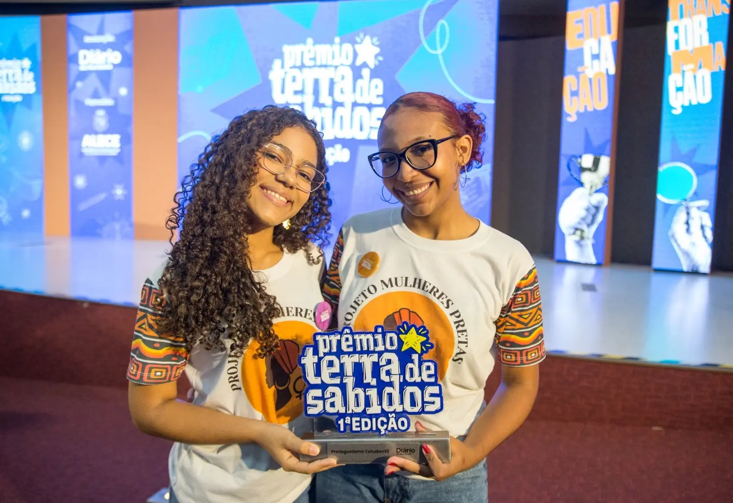Imagem mostra duas adolescentes negras segurando um troféu do Prêmio Terra de Sabidos. À esquerda, uma menina de cabelos cacheados longos. À direita, uma menina de óculos de grau e cabelos ruivos presos em coque.