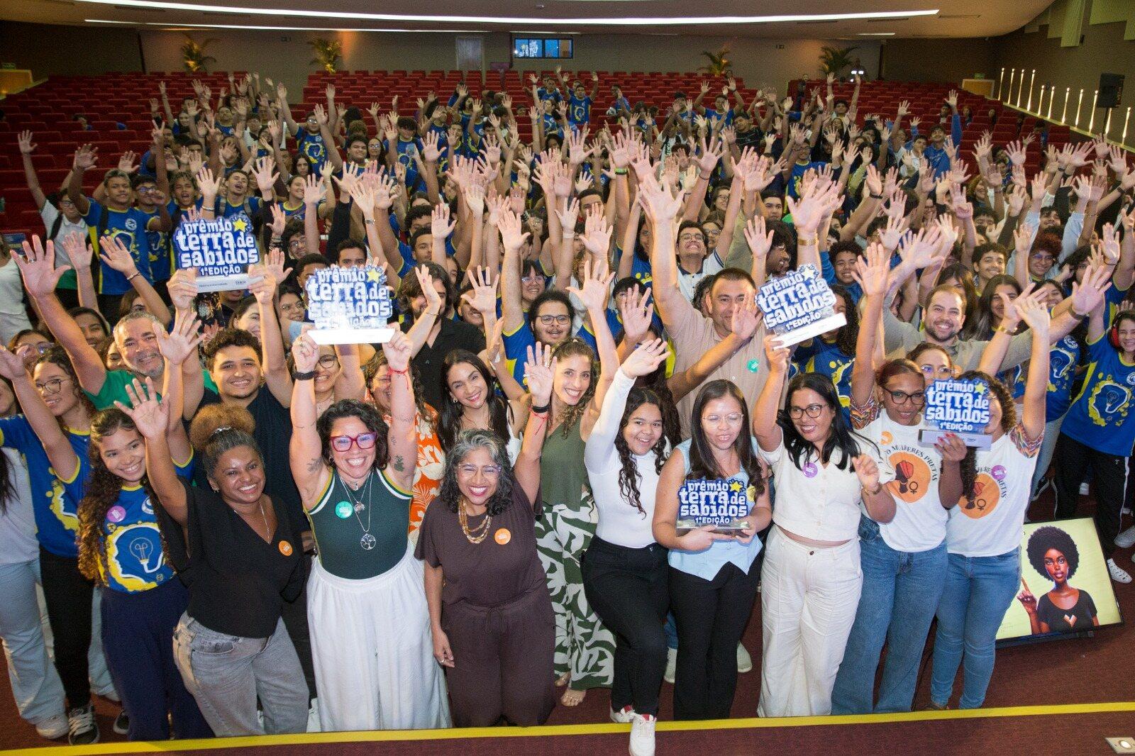 Imagem mostra multidão de alunos e professores em auditório, todos com os braços para cima em comemoração ao Prêmio Terra de Sabidos.