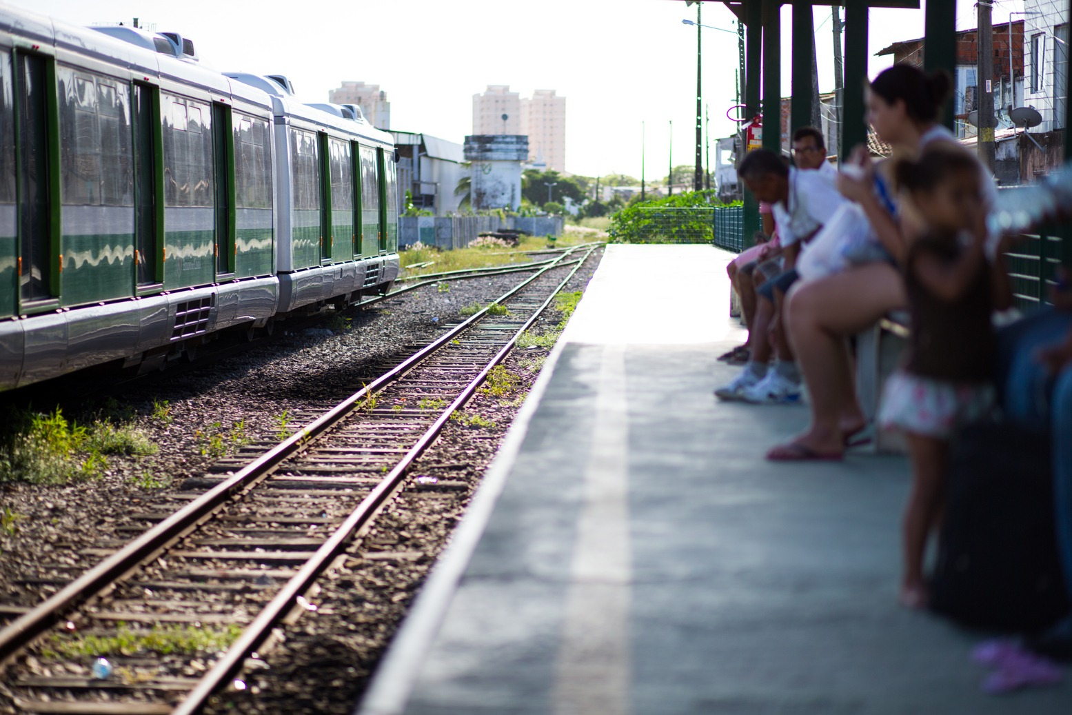 Foto que contém VLT em movimento em estação ferroviária de Fortaleza. Pessoas aguardam sentadas na estação.