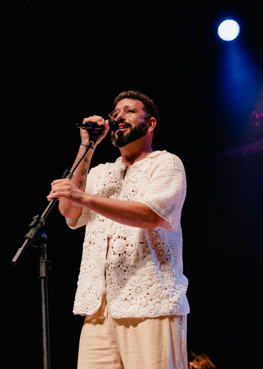 Cantor Juruviara canta em um microfone. Ele tem cabelo, barba e bigode pretos e curtos, usa óculos e camisa e calça de cores claras.