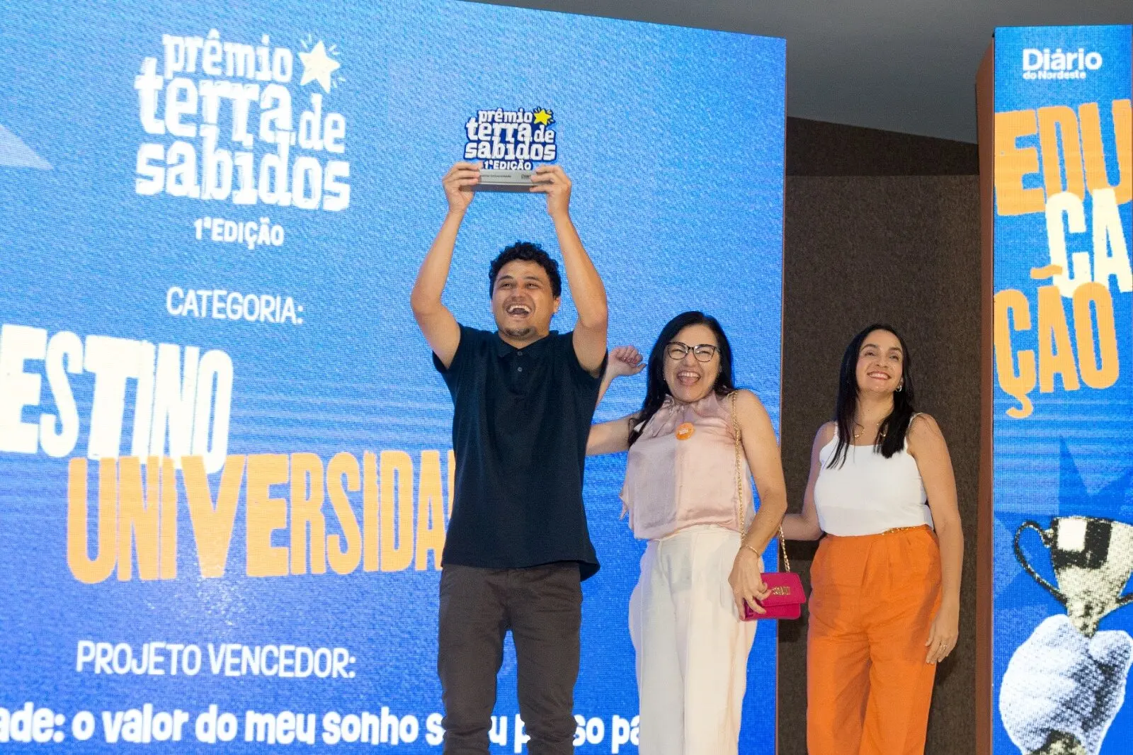 Imagem mostra entrega do Prêmio Terra de Sabidos a três pessoas: um homem vestindo camisa preta e calça cinza ergue o troféu. Ao lado dele, duas mulheres sorriem e comemoram.