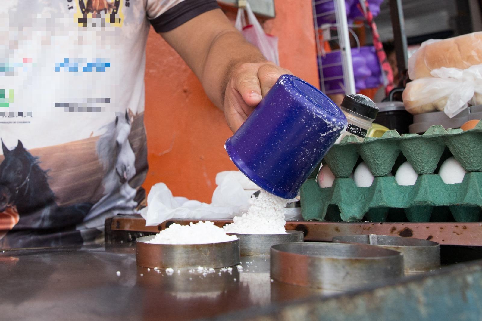 Mão de pessoa branca despejando goma de tapioca branca na chapa, com caixa de ovos em uma bandeja em segundo plano.