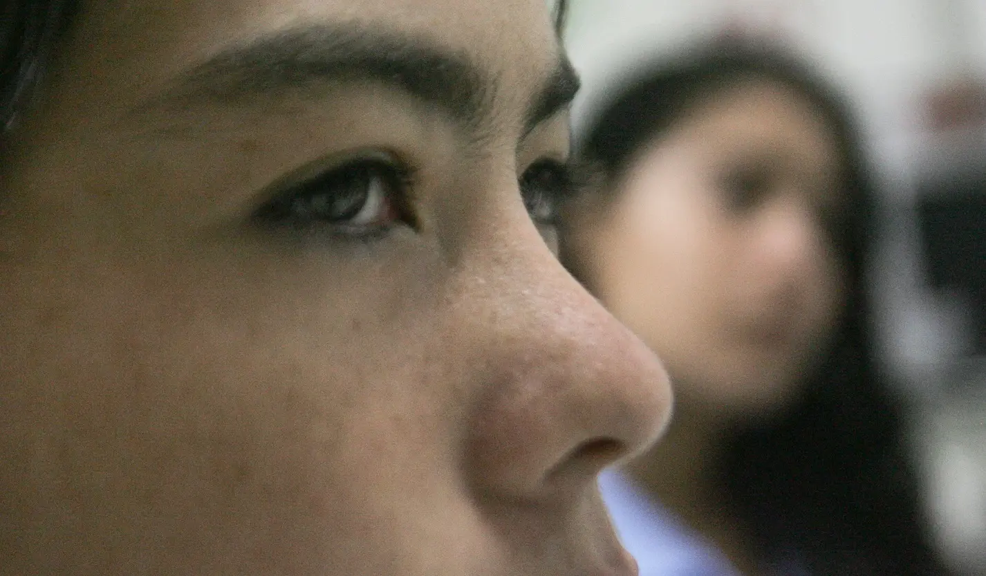 A foto mostra o close do rosto de uma adolescente, com foco em seu olhar sério e concentrado. No fundo desfocado, vê-se outra pessoa. A expressão transmite introspecção, atenção ou preocupação.