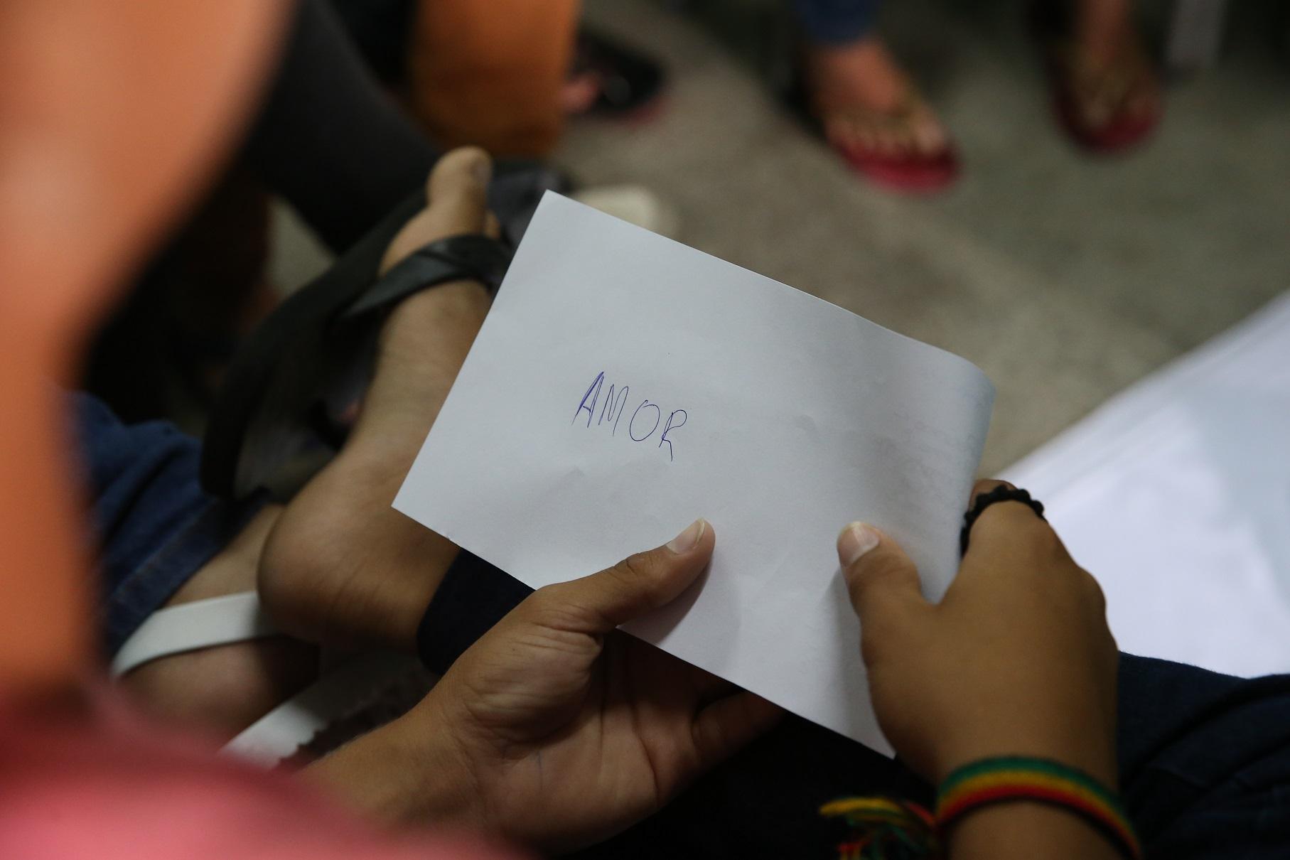 Foto foca as mãos de um jovem segurando um pedaço de papel com a palavra “AMOR” escrita à caneta. A cena ocorre em um ambiente coletivo, possivelmente uma roda de conversa ou atividade educativa.