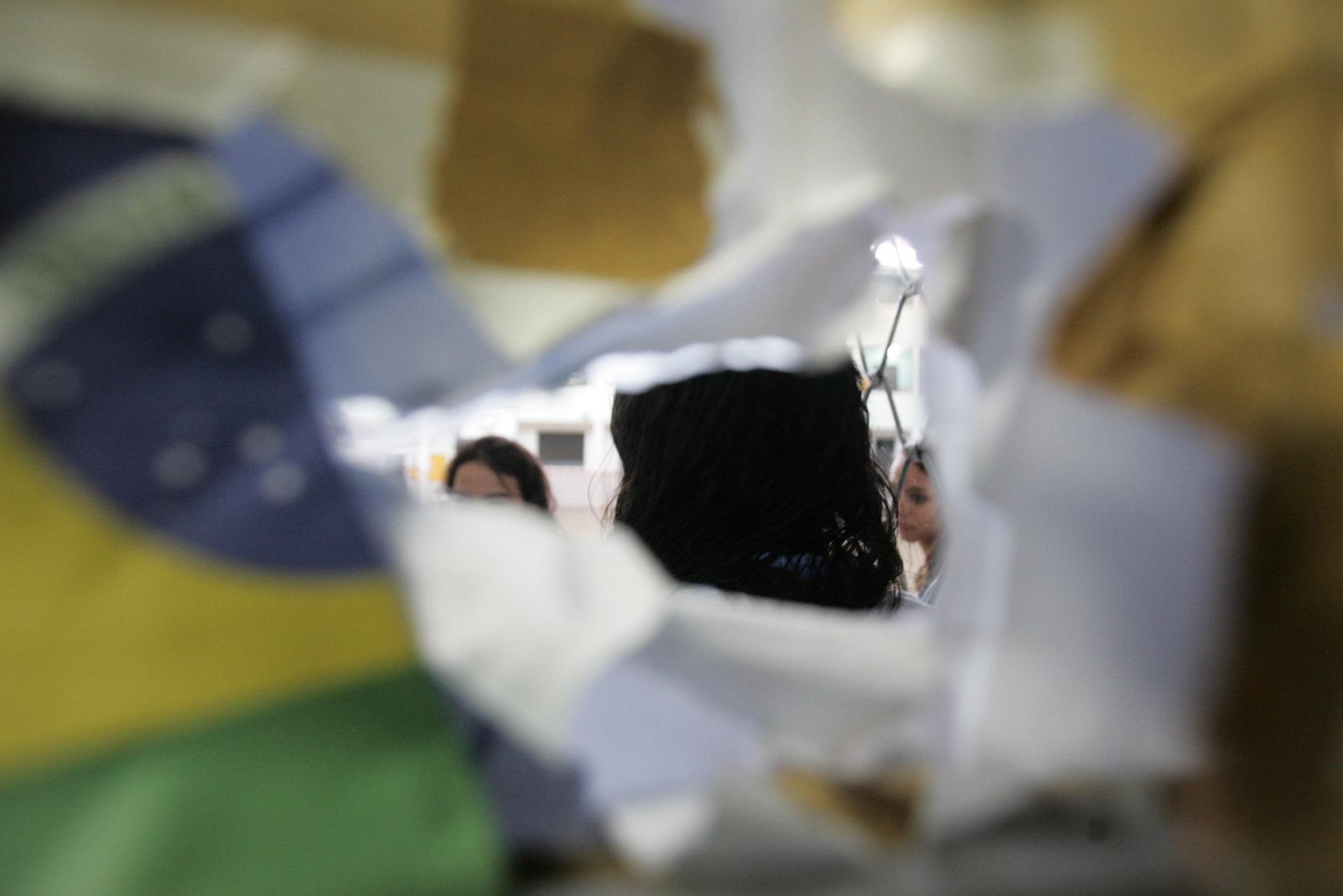 A imagem é feita como se fosse através de um buraco rasgado em papéis com cores variadas (incluindo o padrão da bandeira do Brasil). Atrás, um grupo de pessoas aparece desfocado, sugerindo um cenário de dinâmica escolar em atividade reflexiva.