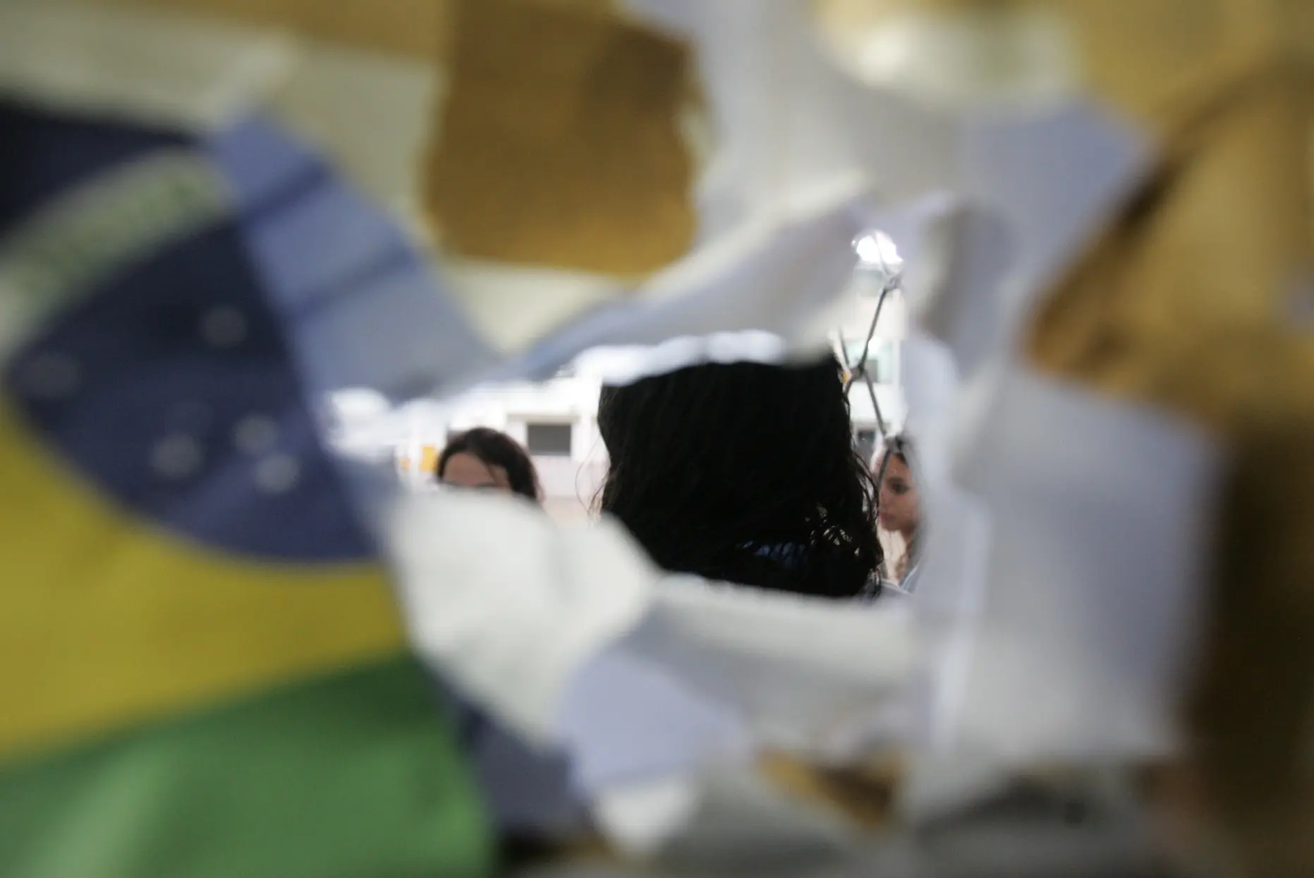 A imagem é feita como se fosse através de um buraco rasgado em papéis com cores variadas (incluindo o padrão da bandeira do Brasil). Atrás, um grupo de pessoas aparece desfocado, sugerindo um cenário de dinâmica escolar em atividade reflexiva.