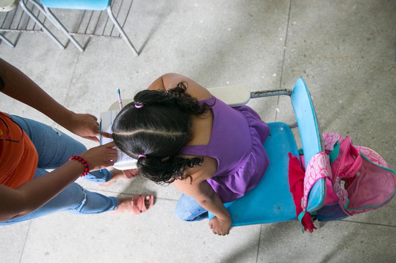 Foto capturada de cima e mostra uma criança pequena sentada em uma carteira escolar azul, com uma mochila rosa pendurada no encosto. Outra pessoa está à sua frente, apontando algo em um caderno. A cena transmite ambiente escolar, aprendizagem e acompanhamento.