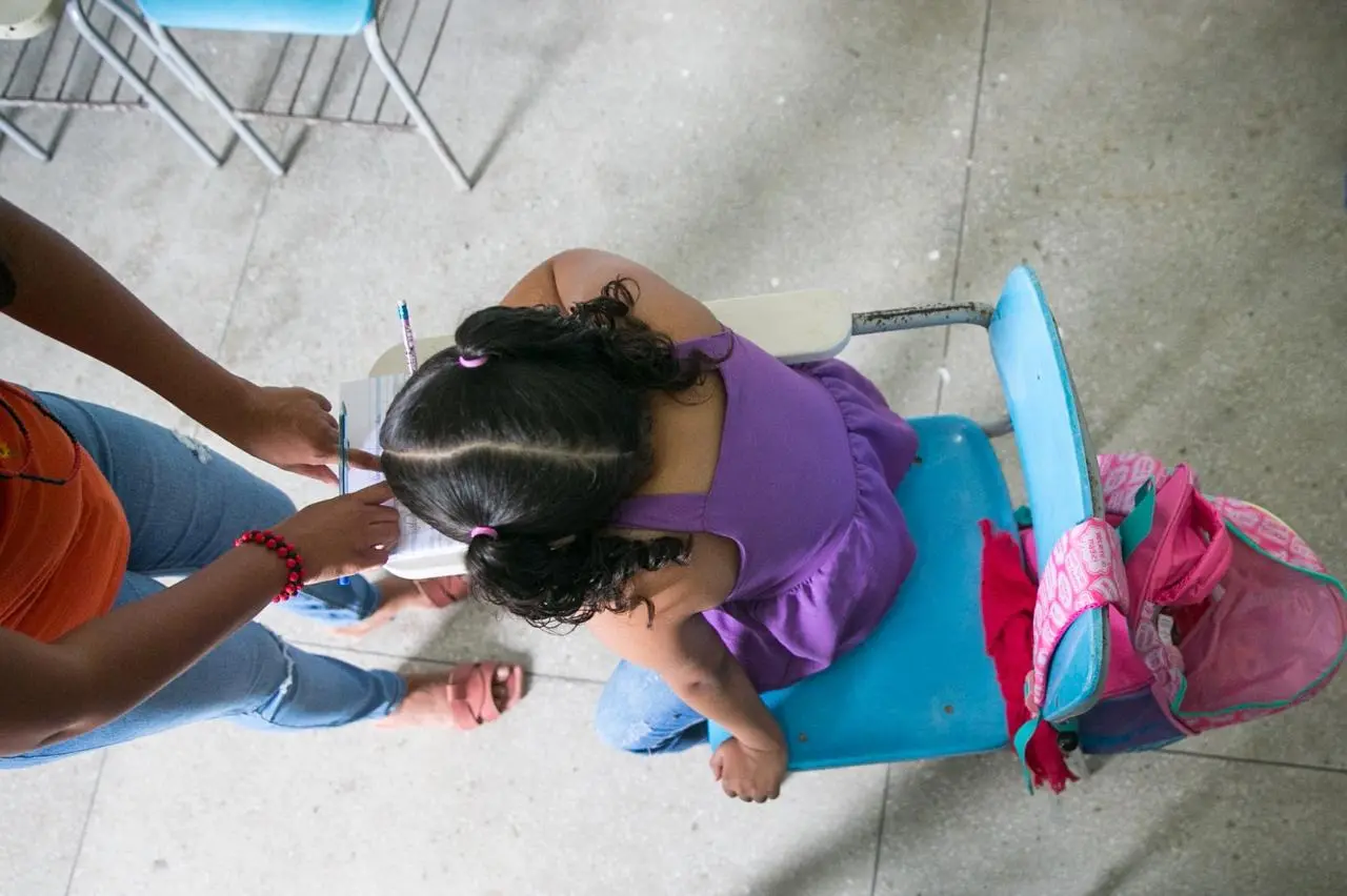 Foto capturada de cima e mostra uma criança pequena sentada em uma carteira escolar azul, com uma mochila rosa pendurada no encosto. Outra pessoa está à sua frente, apontando algo em um caderno. A cena transmite ambiente escolar, aprendizagem e acompanhamento.