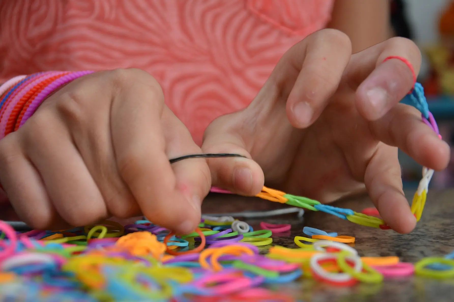 A imagem mostra as mãos de uma criança manipulando pequenos elásticos coloridos, como se estivesse montando pulseiras artesanais. A criança veste uma camisa rosa e usa pulseirinhas no braço. É um momento de atividade manual e criativa.