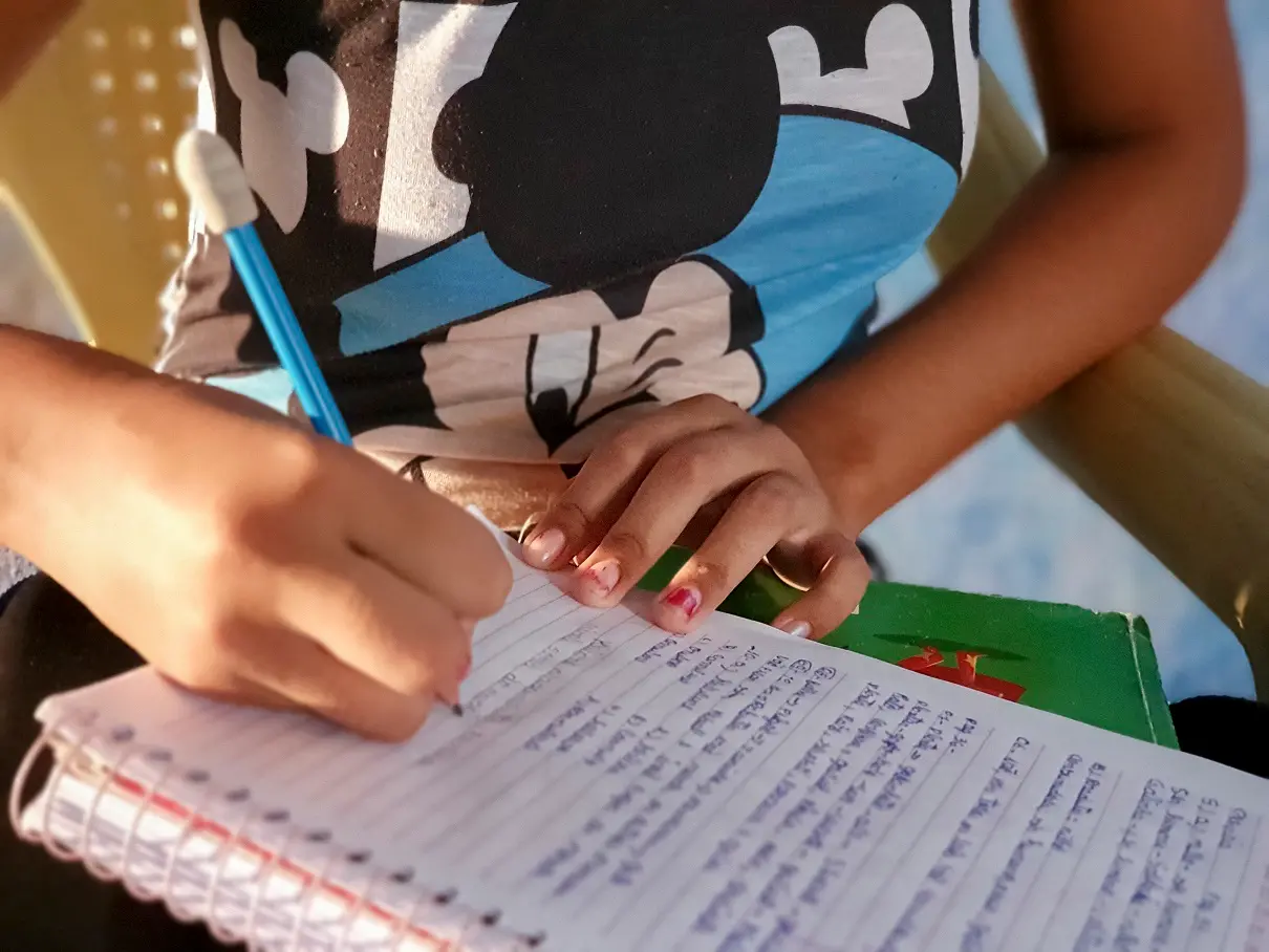 Foto próxima mostra o tronco e as mãos de uma adolescente escrevendo em um caderno que está sobre seu colo. Ela segura um lápis azul e usa uma blusa estampada com personagem infantil. A cena transmite estudo e concentração.