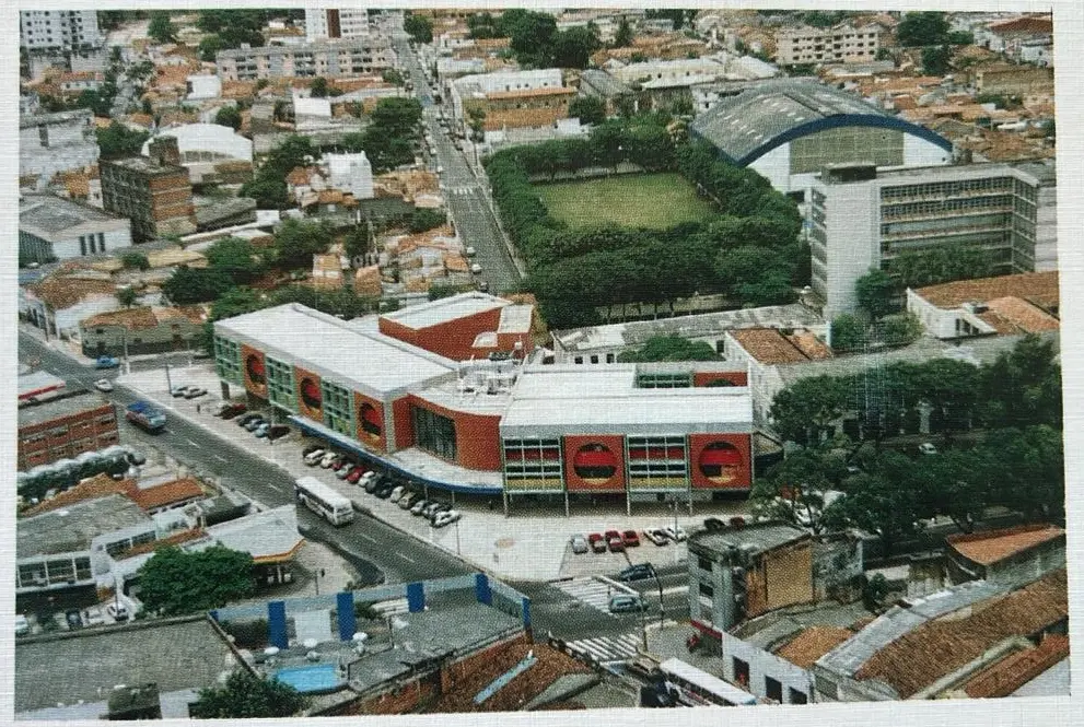 Foto aérea (anos 90/2000) do moderno Colégio Marista, com fachadas vermelhas/brancas e janelas circulares amarelas. Está em área urbana densa. No fundo, há um campo gramado cercado por árvores e um ginásio de teto arqueado, indicando grande complexo educacional.