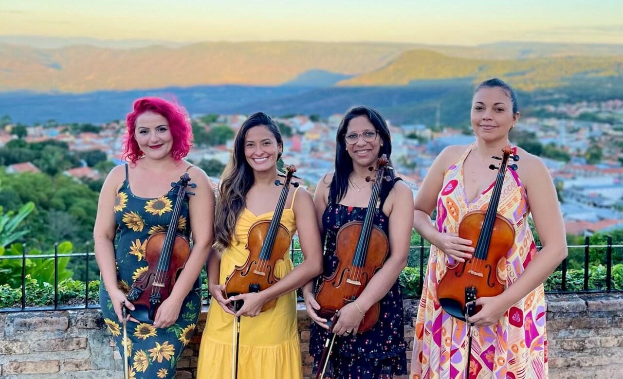 Quatro mulheres jovens sorriem para a foto enquantyo seguram seus instrumentos. Elas usam vestidos coloridos e posam em frente à uma paisagem de serra.