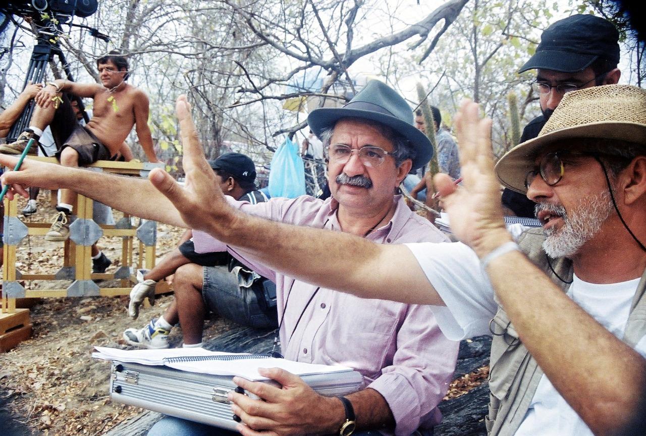 Rosemberg Cariry (de chapéu cinza, no centro) e Antonio Luiz Mendes (de chapéu de palha, à direita) gesticulam em primeiro plano no set de filmagem do filme 'Lua Cambará'. Rosemberg segura um roteiro no colo. Outros membros da equipe e equipamentos de filmagem estão visíveis ao fundo, em um cenário de mata seca.