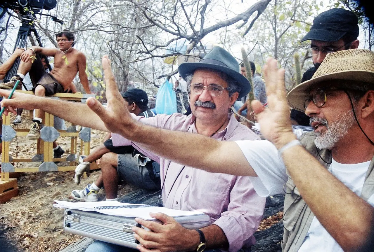 Rosemberg Cariry (de chapéu cinza, no centro) e Antonio Luiz Mendes (de chapéu de palha, à direita) gesticulam em primeiro plano no set de filmagem do filme 'Lua Cambará'. Rosemberg segura um roteiro no colo. Outros membros da equipe e equipamentos de filmagem estão visíveis ao fundo, em um cenário de mata seca.
