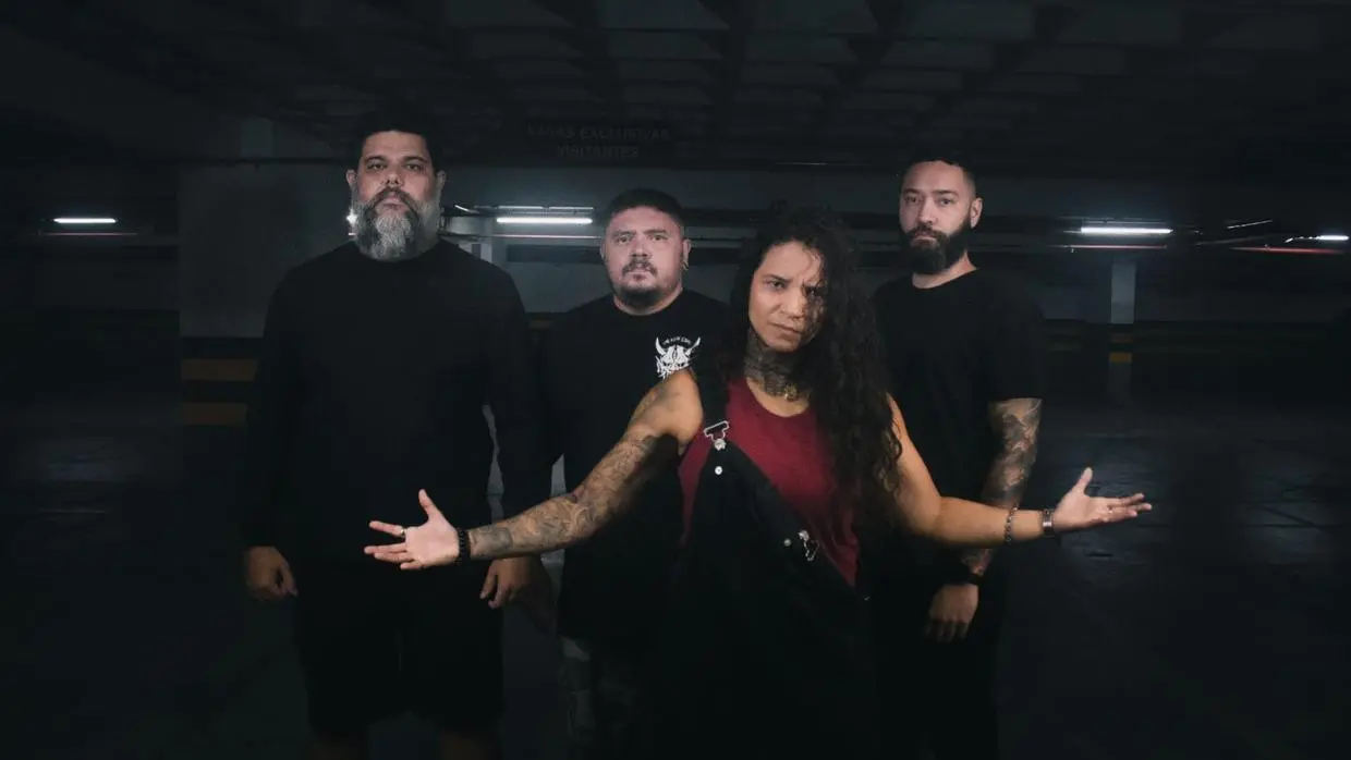 Foto dos membros da banda Corja!, formada por quatro membros: três homens de preto que estão atrás de uma mulher de braços abertos, usando blusa vermelha e macacão preto;