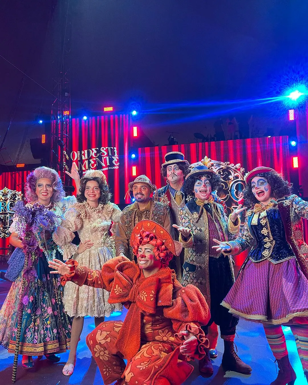 Grupo de seis artistas de circo em figurinos elaborados e coloridos, posando em frente a um palco com iluminação vermelha e azul vibrante. Os figurinos remetem a uma estética circense clássica e teatral. Um artista, com cabelo e roupa laranja-avermelhada, está agachado na frente. Os demais estão de pé, sorrindo e gesticulando para a câmera, com maquiagens características de palhaços ou personagens teatrais.