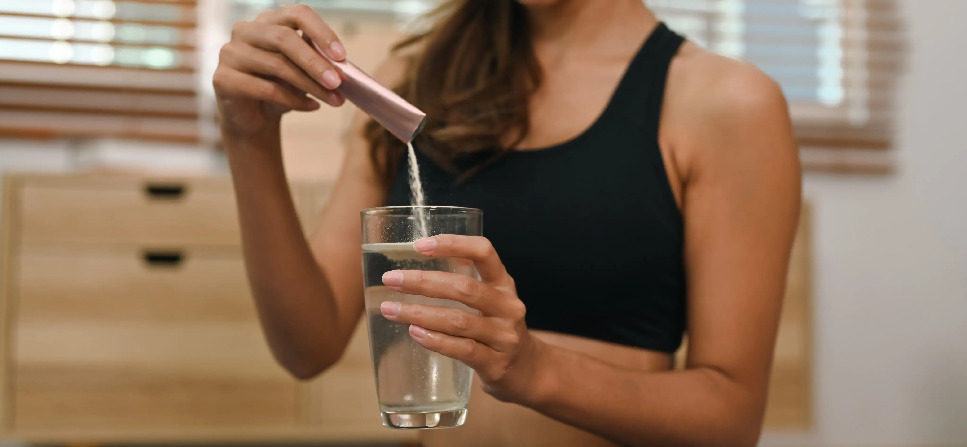 Mulher de top esportivo preto dissolvendo um pó de suplemento (de um sachê) em um copo de água.