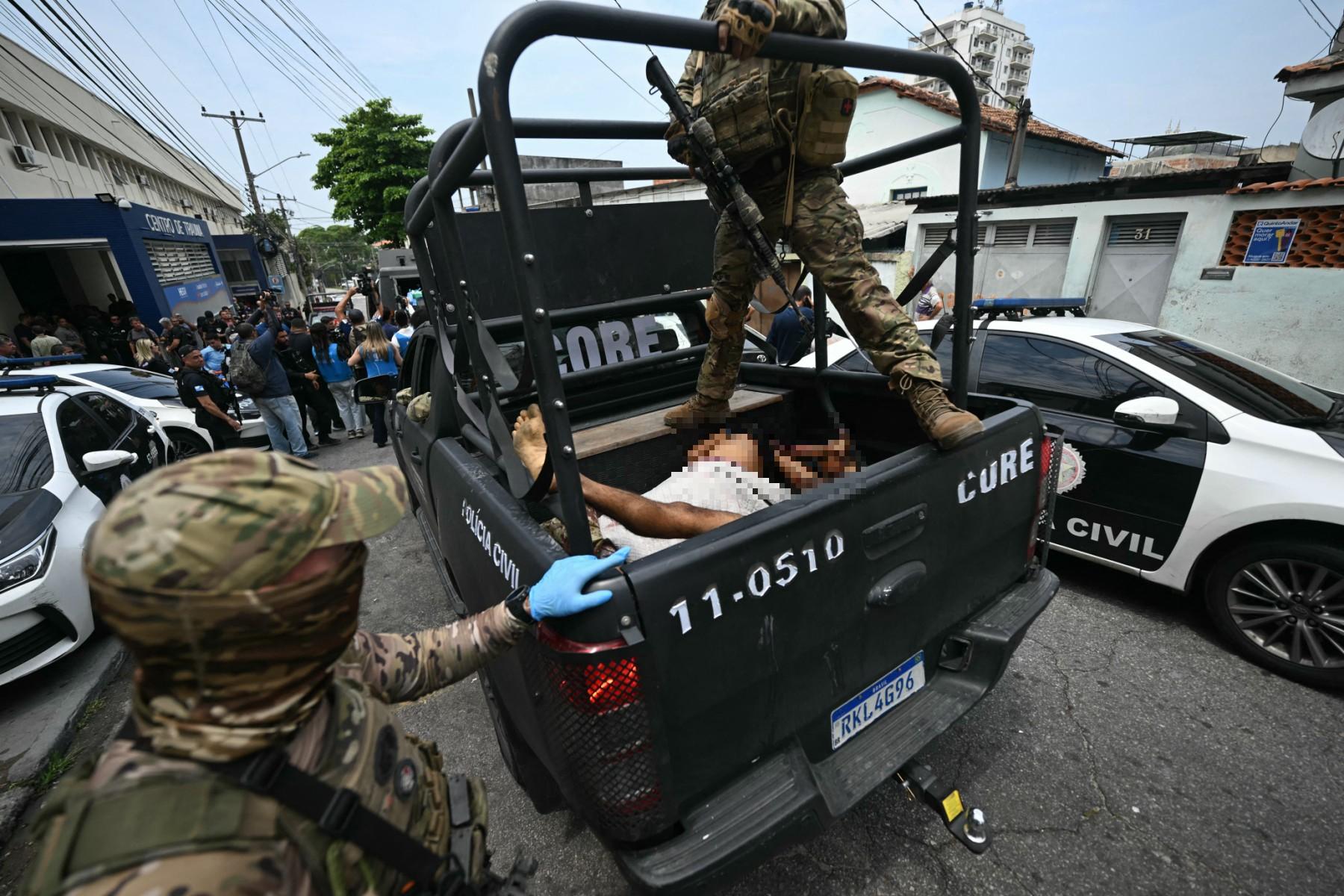 A imagem mostra uma operação policial em uma área urbana. Na cena, agentes fortemente armados e vestidos com uniformes camuflados da Polícia Civil transportam, na caçamba de uma caminhonete tática, corpos cobertos parcialmente por panos. Ao fundo, há outras viaturas, policiais e pessoas reunidas em frente a um prédio identificado como “Sede da Core”, sigla para Coordenadoria de Recursos Especiais. A cena retrata o momento posterior a uma ação policial de grande porte, com forte presença das forças de segurança e grande atenção da imprensa.