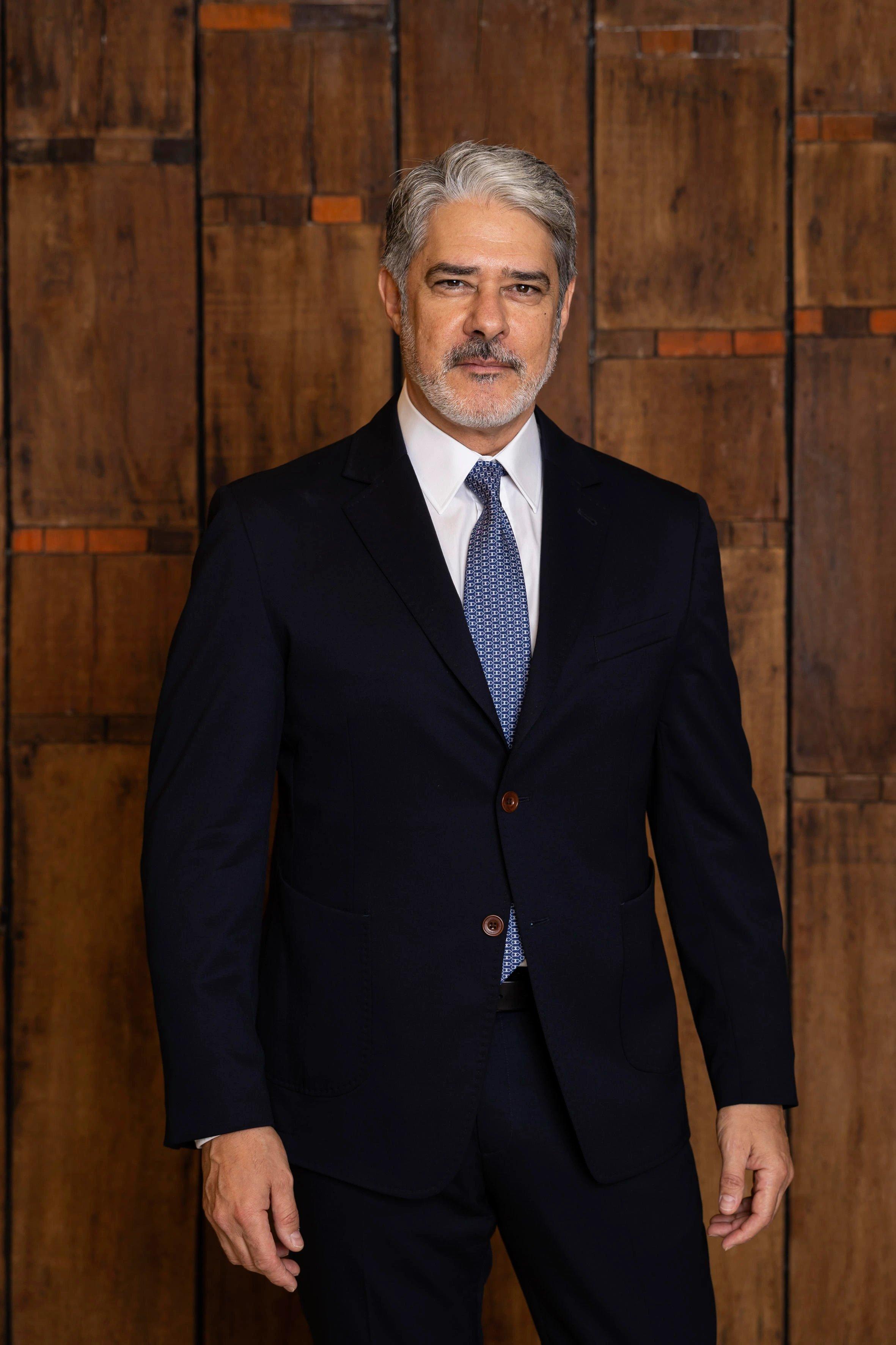 Imagem do apresentador William Bonner com terno escuro e fundo de madeira usada para matéria sobre os bastidores da transição na Globo.