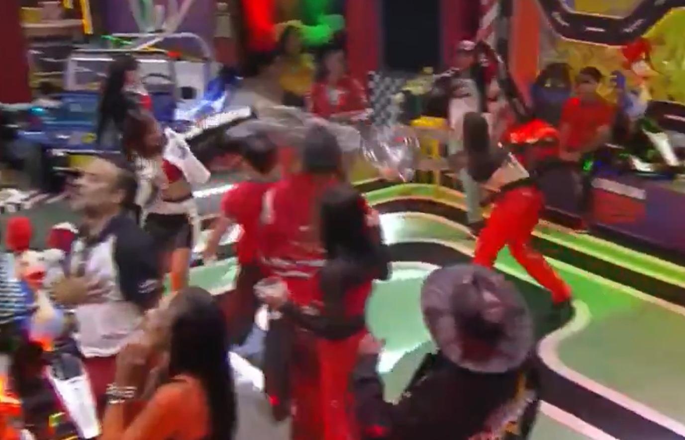 Print de um vídeo do reality show A Fazenda, numa festa colorida temática de Corrida Maluca. Enquanto participantes dançam, uma mulher atira o conteúdo de um balde de gelo em outra, que estende um braço para se defender.