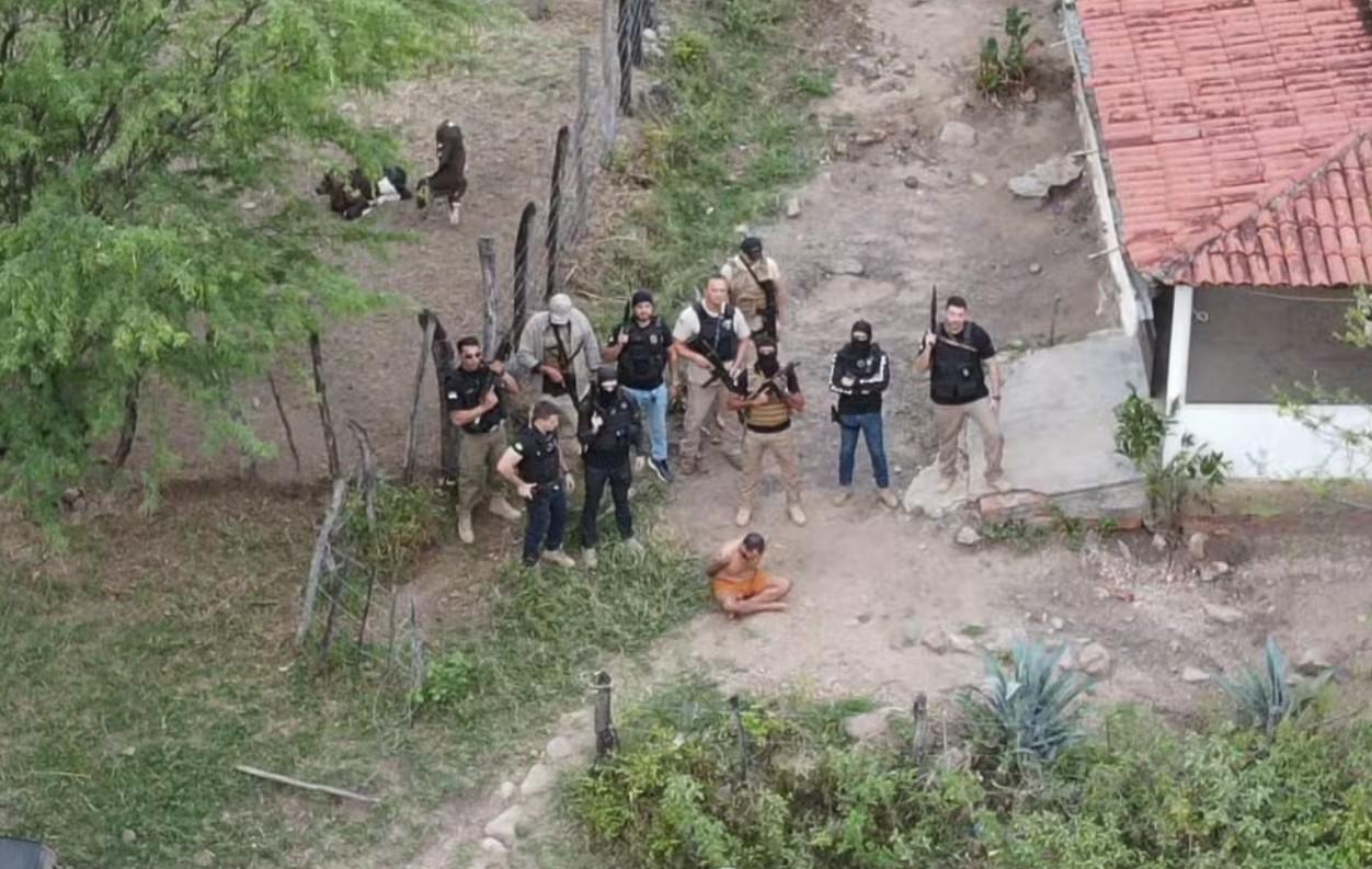 Foto aérea mostra policiais civis fardados e armados posando para câmera enquanto há um homem algemado e sentado no chão, sem camisa. O ambiente é uma área rural com casa de telhas, chão de barro, cerca de arame e algumas árvores.