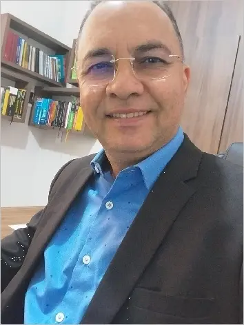 Selfie de José Nanda Bezerra, um advogado calvo, de óculos e usando terno e blusa social azul. Ele sorri para a foto. Ao fundo, há uma estante com vários livros dentro de seu escritório.