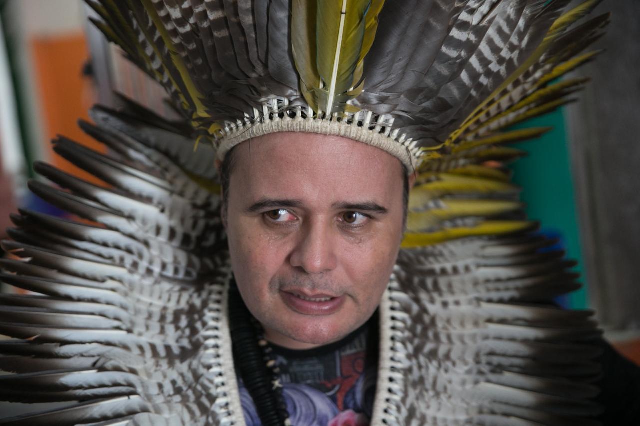 Foto aproximada do rosto de Paulo Anacé, uma líder indígena cearense. Ele usa uma blusa colorida e na cabeça utiliza um grande cocar, cheio de penas brancas e amarelas, que caem em volta de seu pescoço.