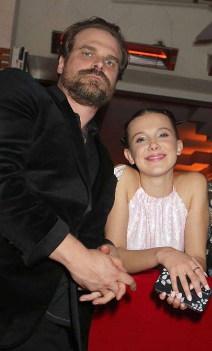 David Harbour, vestindo roupa preta,  e Millie, de vestido branco e segurando uma bolsa de festa.