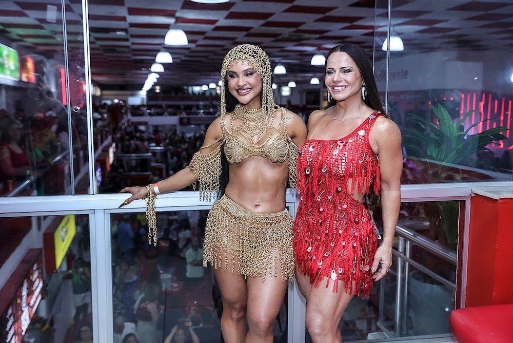 Mileide Mihaile, com figurino de pedrarias douradas, posa para foto ao lado de Viviane Araújo, que usa vestido de franjas vermelhas.