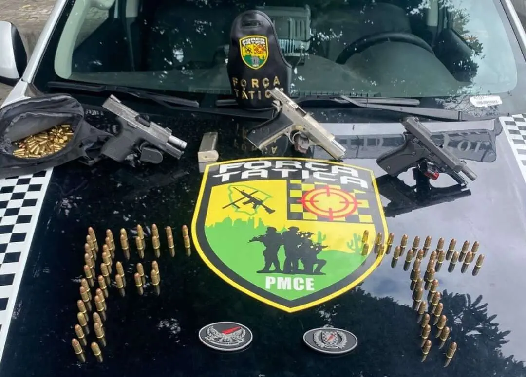 A imagem mostra três armas de fogo e munições em cima de um capô de uma viatura policial.