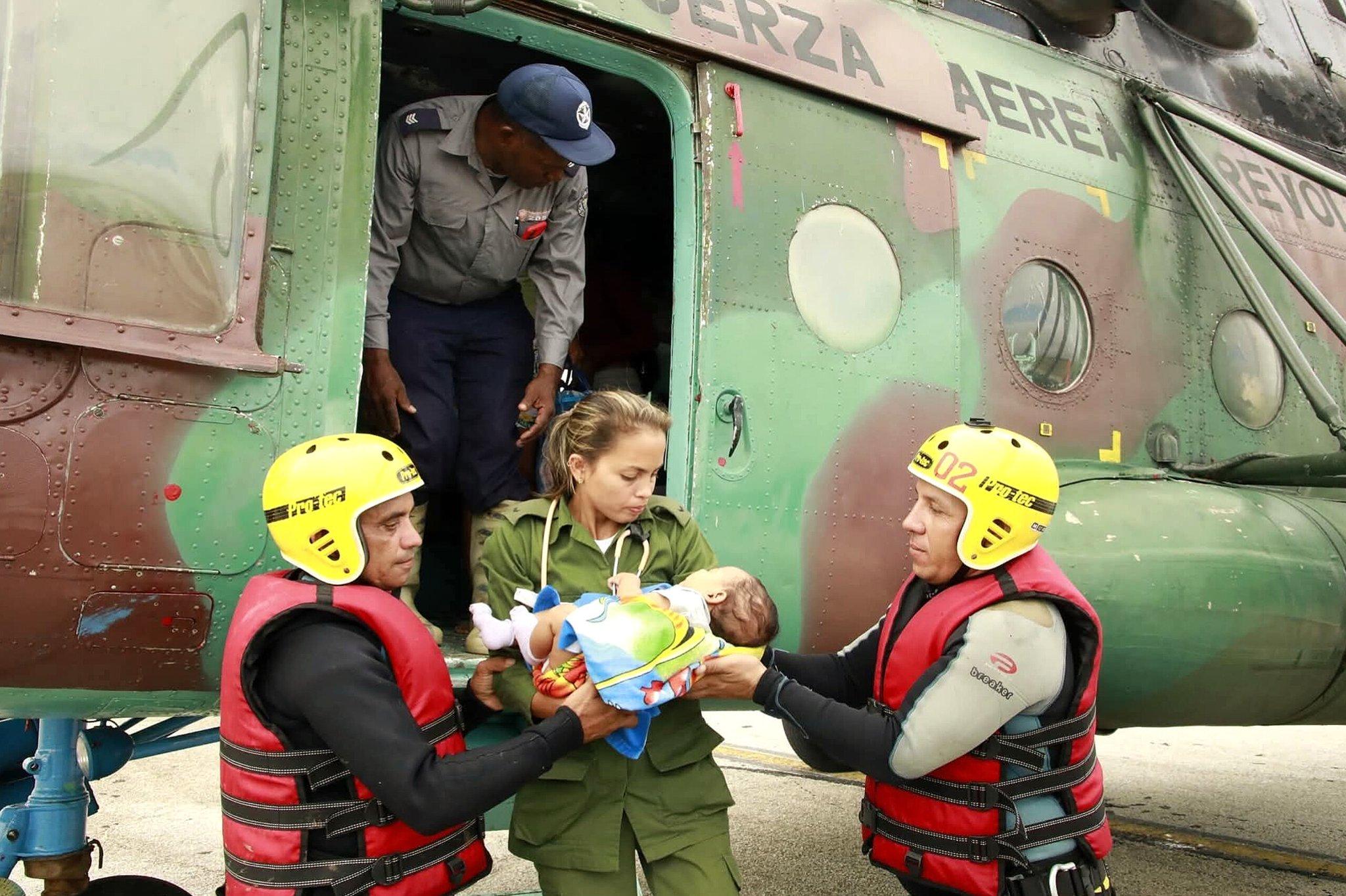 Helicóptero militar camuflado com a porta aberta, onde um integrante da tripulação está próximo à saída. Dois socorristas com capacetes amarelos e coletes vermelhos recebem um bebê enrolado em um cobertor colorido das mãos de uma pessoa vestida com uniforme verde, em uma operação de resgate.