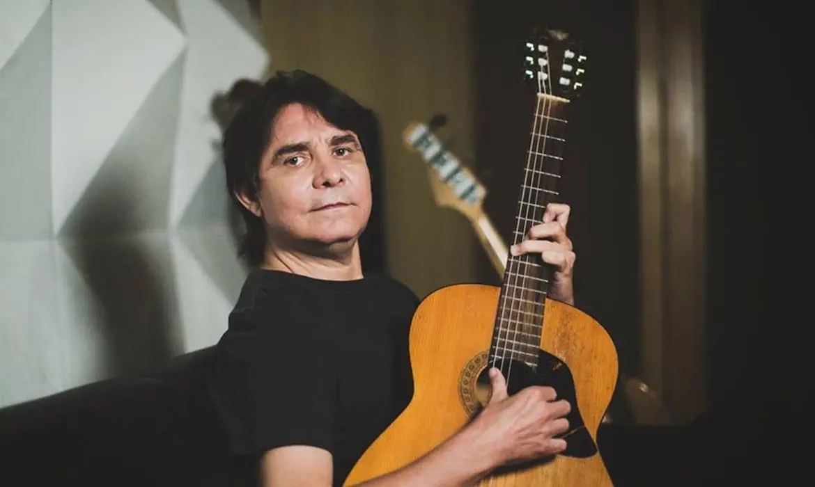Foto do compositor e músico Lo Borges. Ele está sentado, veste camiseta preta lisa, segura um violão, tem cabelos pretos. Está com semblante sério.