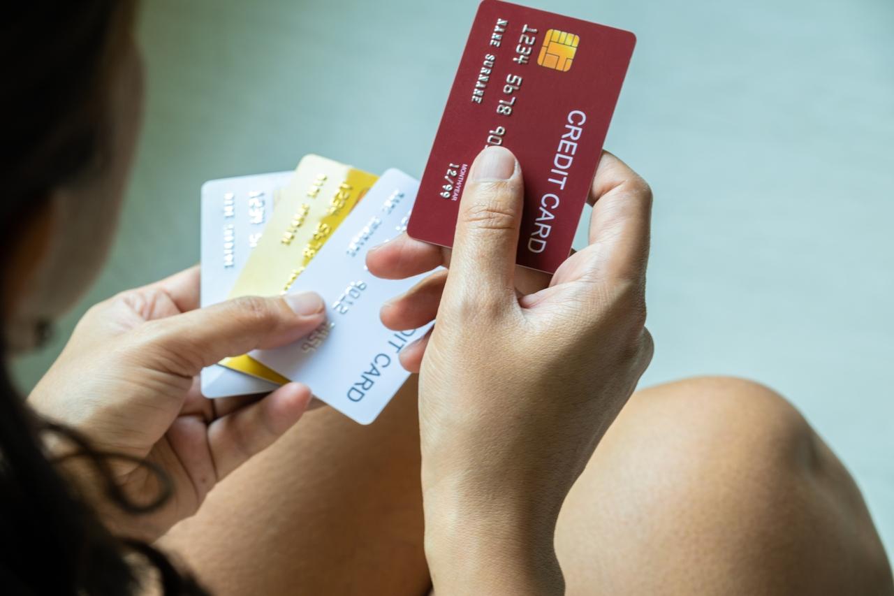 Imagem mostra mãos segurando múltiplos cartões de crédito de diferentes cores, incluindo um vermelho e dourado, ilustrando dívidas financeiras.