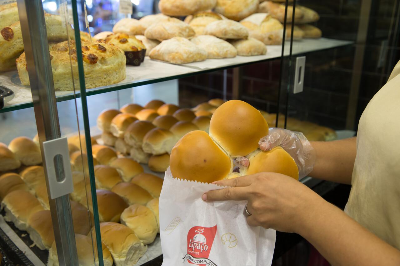 Três pães redondos sendo colocados em saco branco com detalhes de logo vermelho. Ao fundo, pães expostos em duas prateleiras.