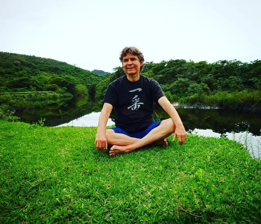 Imagem mostra o cantor e compositor Lô Borges sentado em posição de meditação na grama verde, ao lado de um lago cercado por floresta.