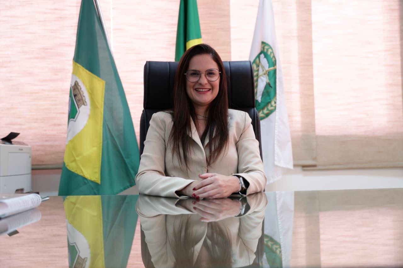 Mulher sentada atrás de uma mesa de vidro, sorrindo, com as mãos apoiadas à frente. Trata-se de Sâmia Farias. Ela usa óculos e blazer claro. Atrás dela há três bandeiras — uma verde e amarela, uma verde e outra branca com o emblema da Defensoria Pública do Estado do Ceará — e uma cortina bege cobrindo a janela.