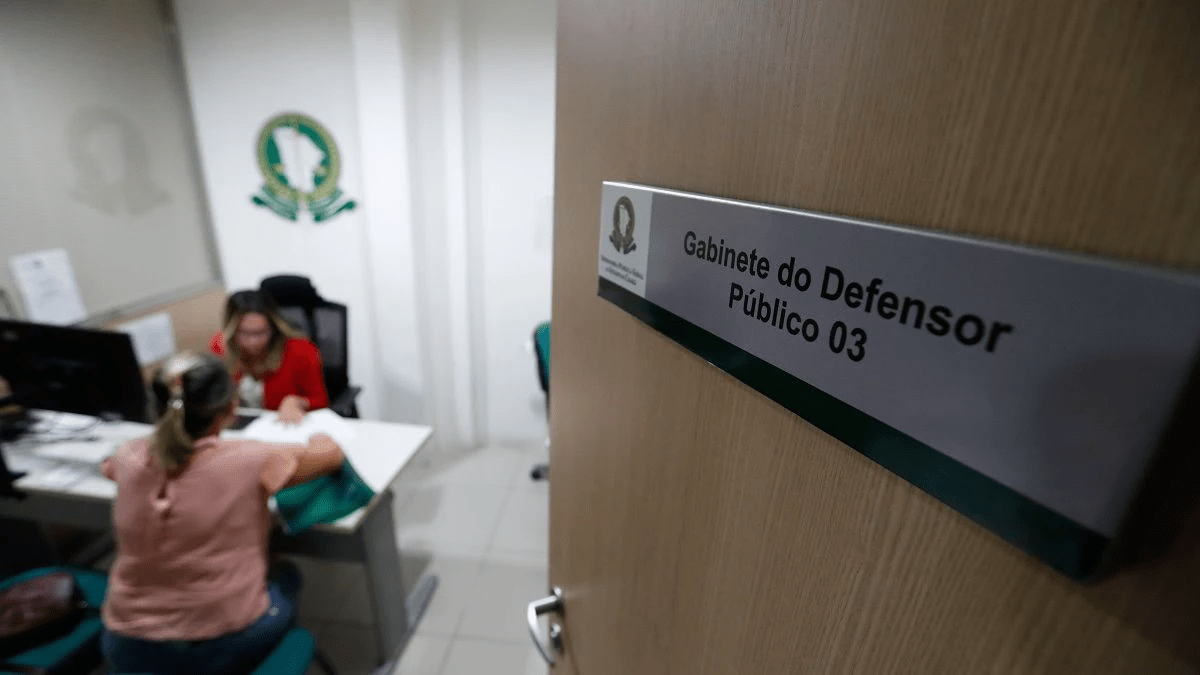 Placa na porta com o texto “Gabinete do Defensor Público 03”. Ao fundo, dentro da sala, uma mulher de roupa vermelha atende outra mulher sentada à frente da mesa. O ambiente tem paredes claras, logotipo da Defensoria Pública do Estado do Ceará na parede e computadores sobre a mesa.