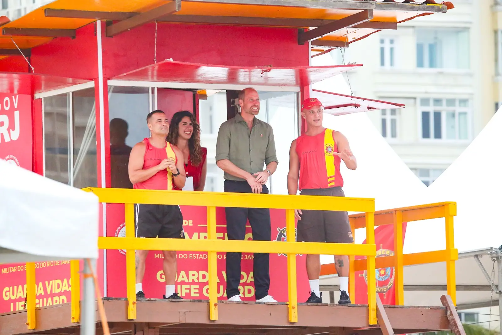 A foto mostra o príncipe William em uma torre de salva-vidas no Rio de Janeiro, acompanhado por dois guarda-vidas e uma mulher. Ele sorri enquanto observa algo à distância, em um momento descontraído durante sua visita oficial à cidade.