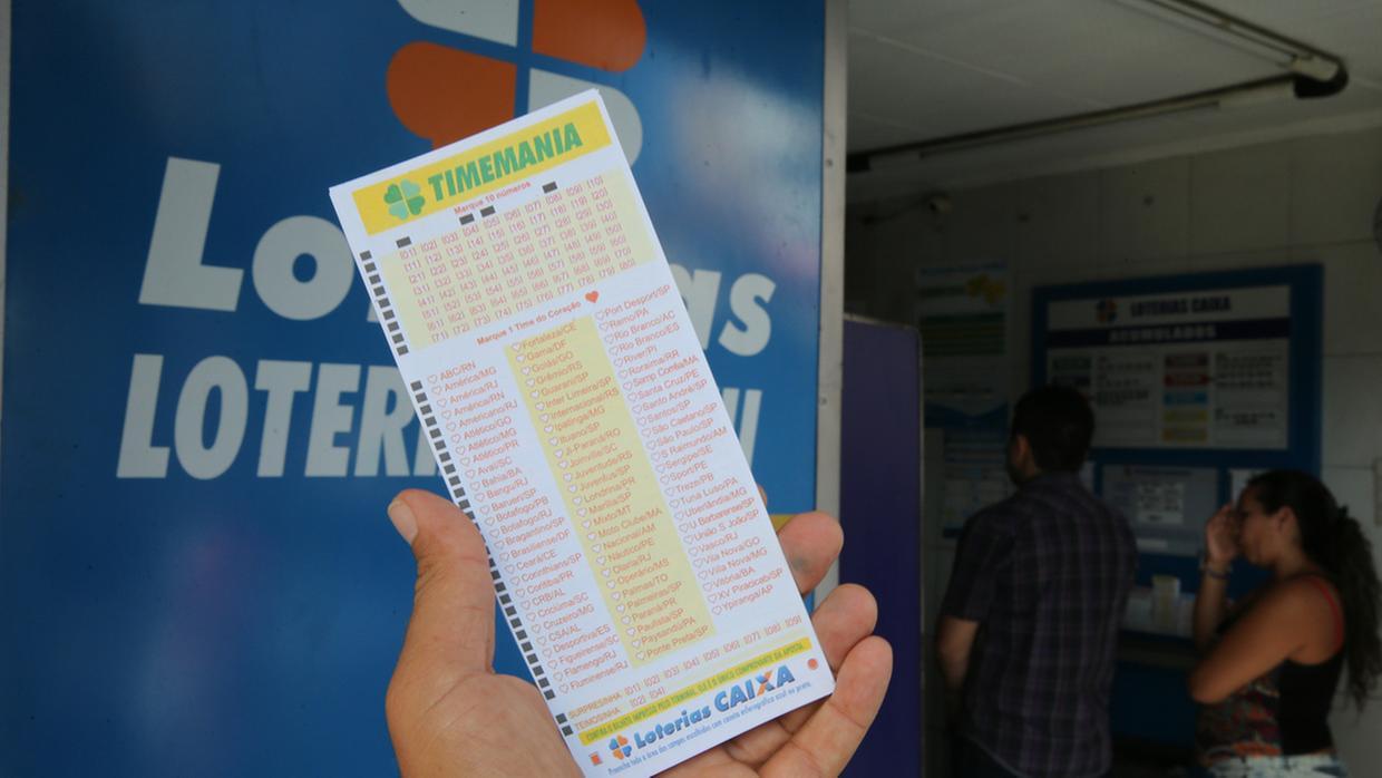 Imagem mostra pessoa segurando um bilhete de loteria ao lado de um caixa e uma urna de apostas oferecendo serviços de loteria da Loterias CAIXA.