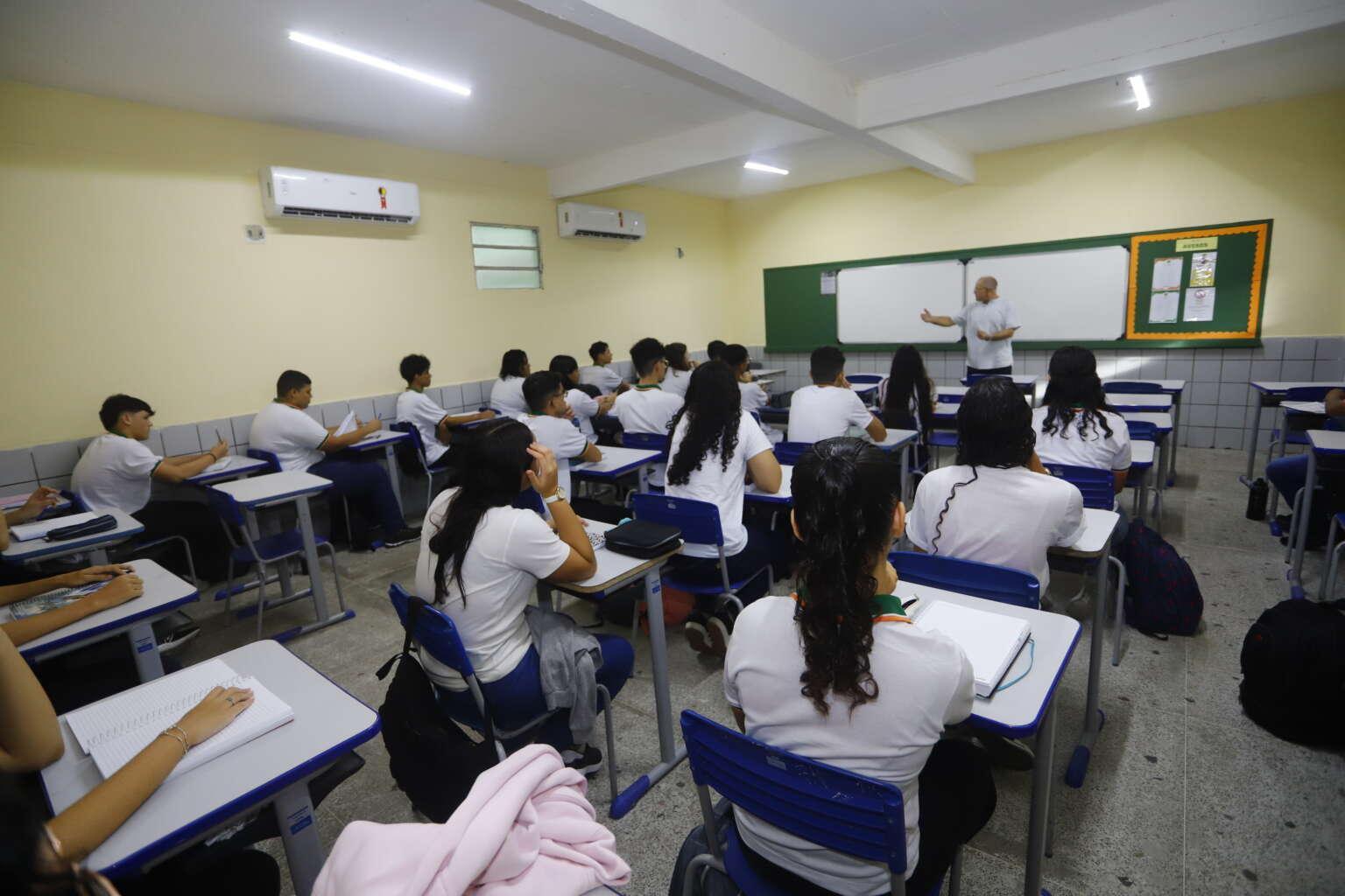 Imagem mostra sala de aula com diversos alunos sentados em suas carteiras, atentos a um professor que gesticula em frente ao quadro branco.