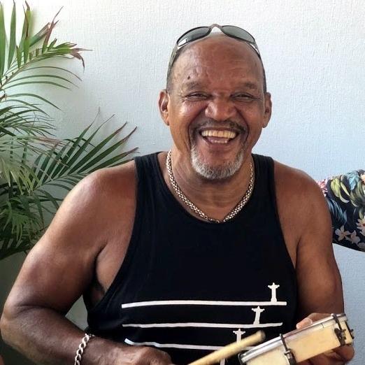 Foto em close de Jorge do Pagode, um homem negro, mais velho, sorrindo amplamente e olhando diretamente para frente. Ele usa óculos de sol na cabeça e uma regata preta com um desenho branco estilizado que parece incluir a silhueta do Cristo Redentor. Ele segura um instrumento de percussão, provavelmente um pandeiro ou tamborim, e uma baqueta.