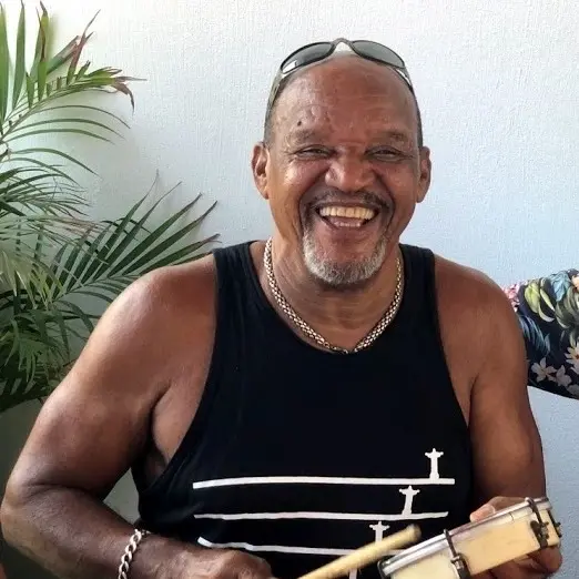 Foto em close de Jorge do Pagode, um homem negro, mais velho, sorrindo amplamente e olhando diretamente para frente. Ele usa óculos de sol na cabeça e uma regata preta com um desenho branco estilizado que parece incluir a silhueta do Cristo Redentor. Ele segura um instrumento de percussão, provavelmente um pandeiro ou tamborim, e uma baqueta.