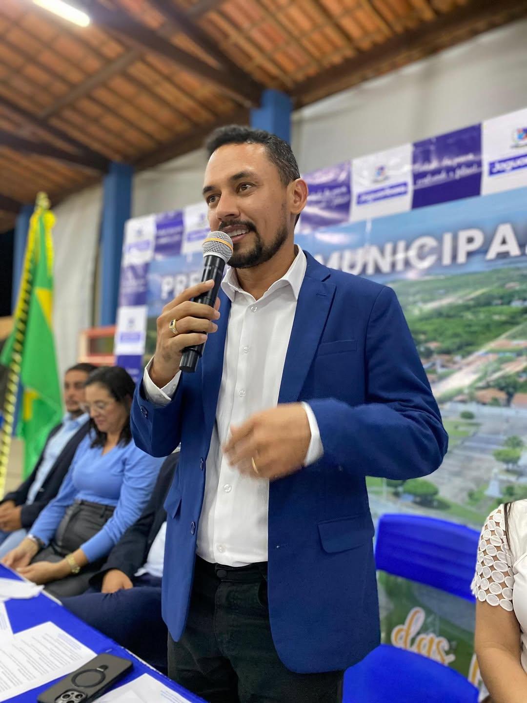 O prefeito de Jaguaribara está falando ao microfone em um evento oficial. Ele veste paletó azul e camisa branca, e está em pé diante de uma mesa coberta por toalha azul, onde há papéis e um celular. Atrás dele, um painel traz o nome da prefeitura e a bandeira do Brasil está posicionada ao fundo. Outras pessoas estão sentadas na mesa, ouvindo a fala. O ambiente parece ser um ginásio coberto, com teto de telhas e iluminação amarela.