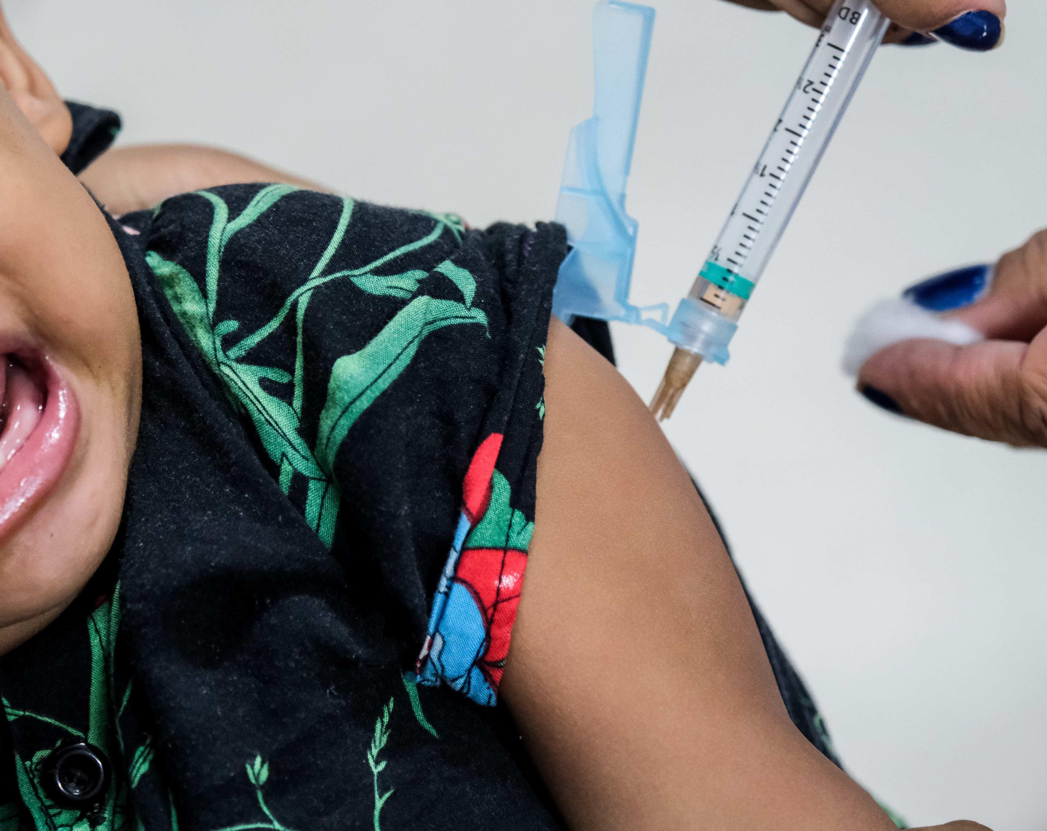 Close-up de um bebê chorando enquanto um profissional de saúde administra uma vacina em seu braço direito com uma seringa.