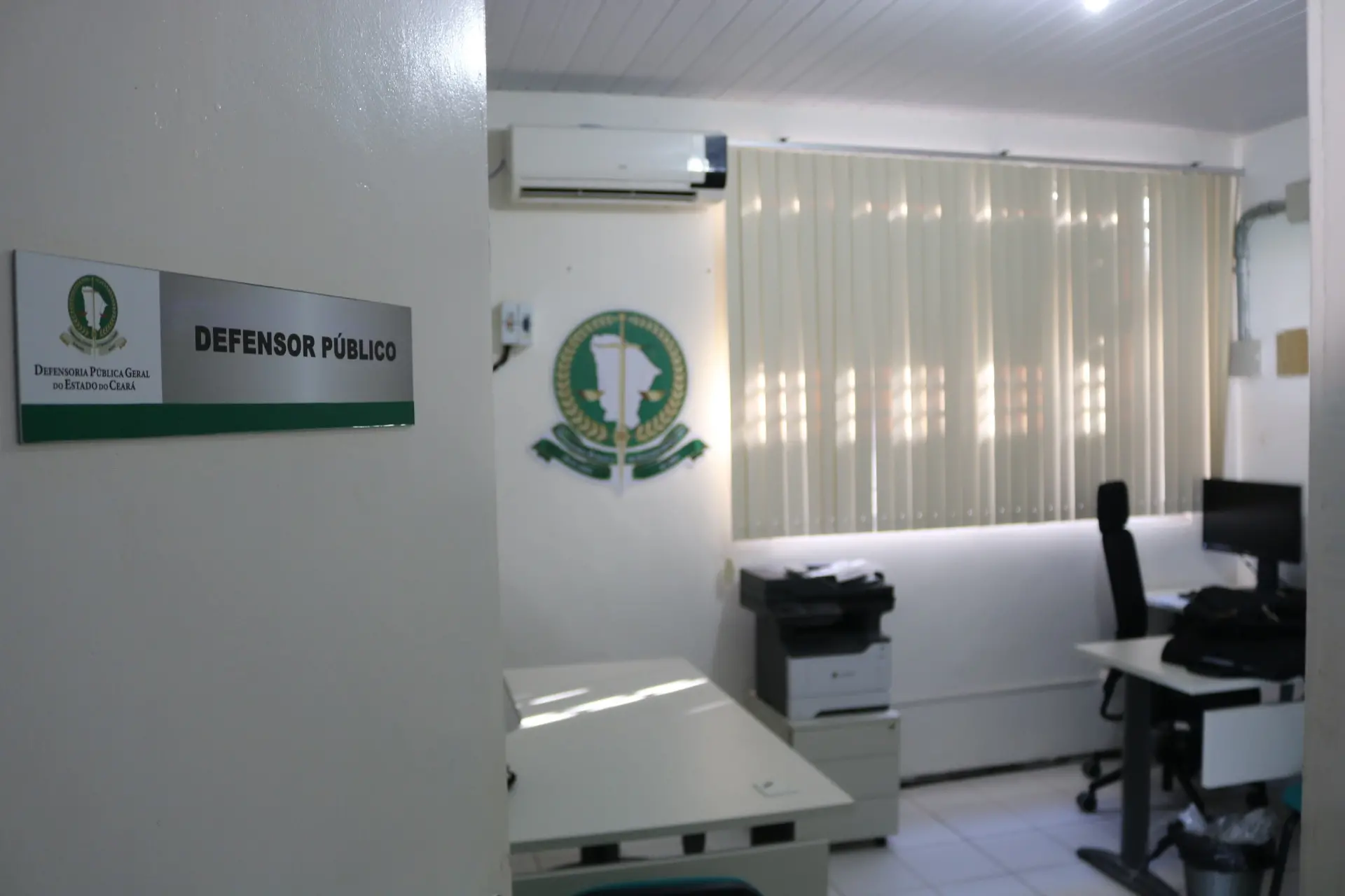Sala de atendimento da Defensoria Pública Geral do Estado do Ceará. Na porta, há uma placa com o logotipo da instituição e o texto “Defensor Público”. O ambiente interno é iluminado, com paredes brancas, cortinas verticais bege cobrindo a janela e o brasão da Defensoria fixado na parede. Há mesas com cadeiras, computador, impressora e outros equipamentos de escritório organizados no espaço.