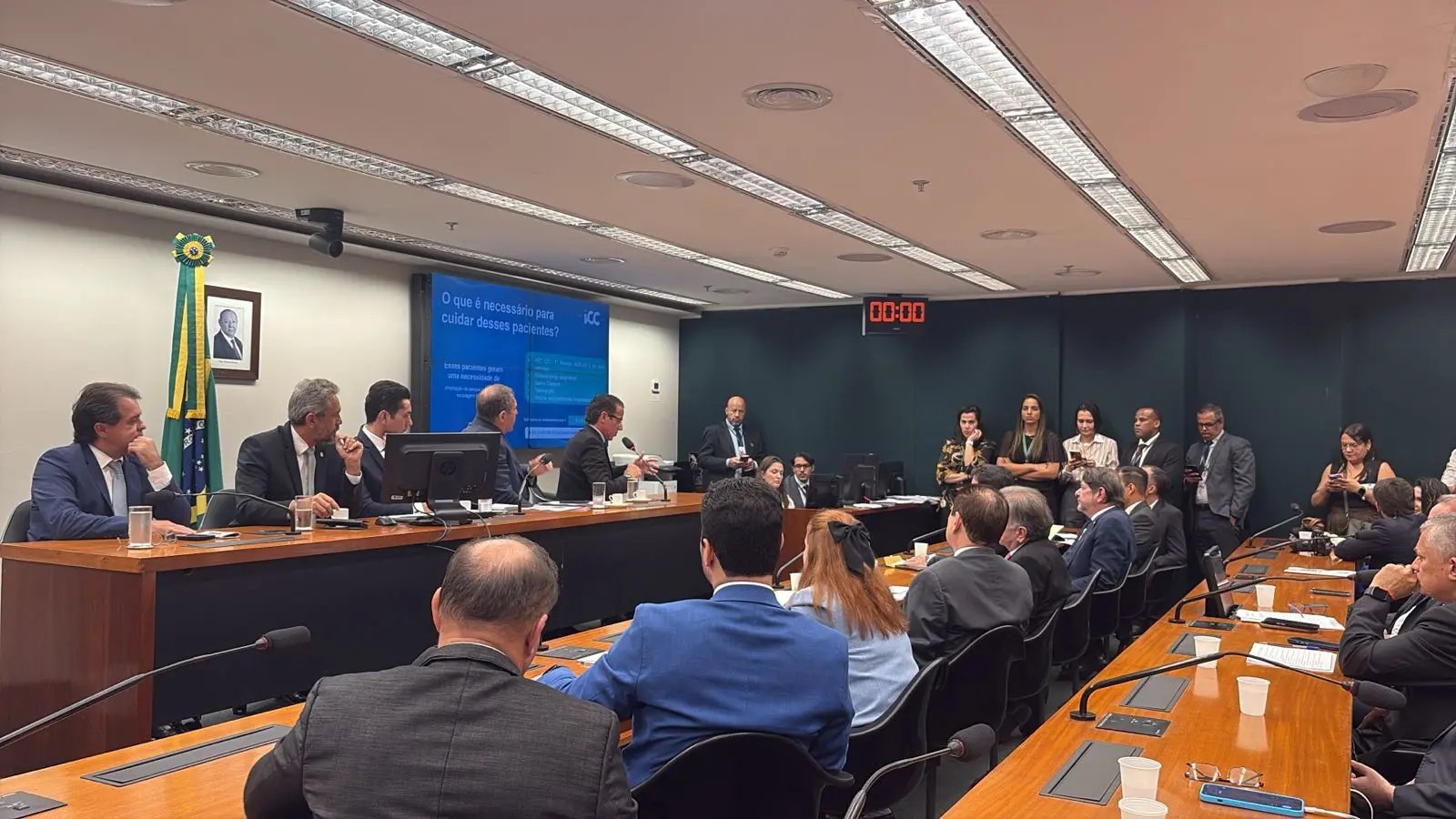 Em uma sala de comissão da Câmara dos Deputados, o governador do Ceará, Elmano de Freitas, e o deputado federal Evandro Leitão participam de uma reunião ao lado de outros parlamentares. Eles estão sentados à mesa principal, com microfones, copos d’água e papéis à frente. Ao fundo, há uma tela com a apresentação “ICC como solução para esse problema” e a bandeira do Brasil. A cena mostra um ambiente formal de debate legislativo, com assessores e técnicos acompanhando a sessão.